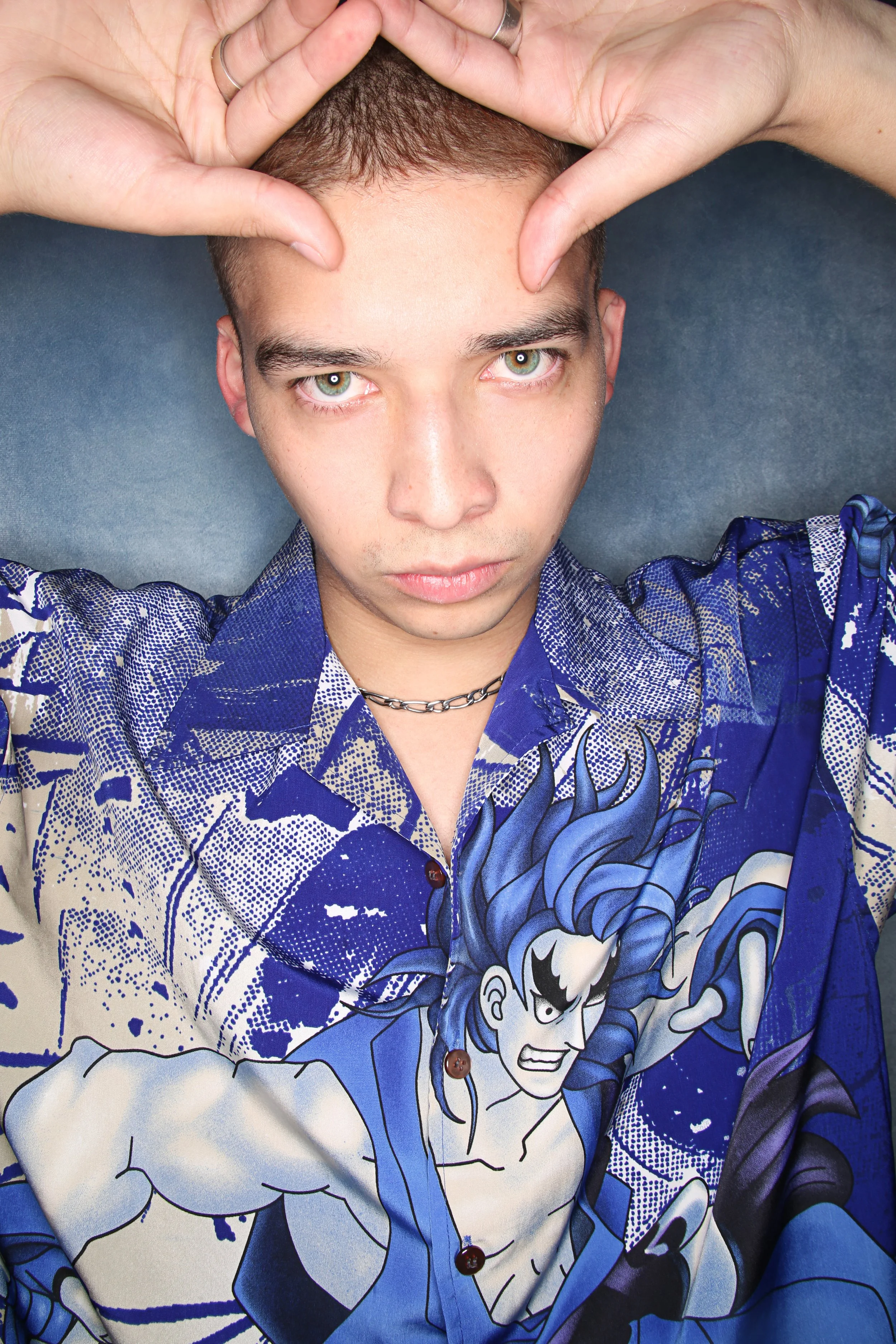 A person with short brown hair and green eyes lying on a blue surface, wearing a colorful blue and white patterned shirt featuring comic book characters and designs, with hands resting on their forehead.