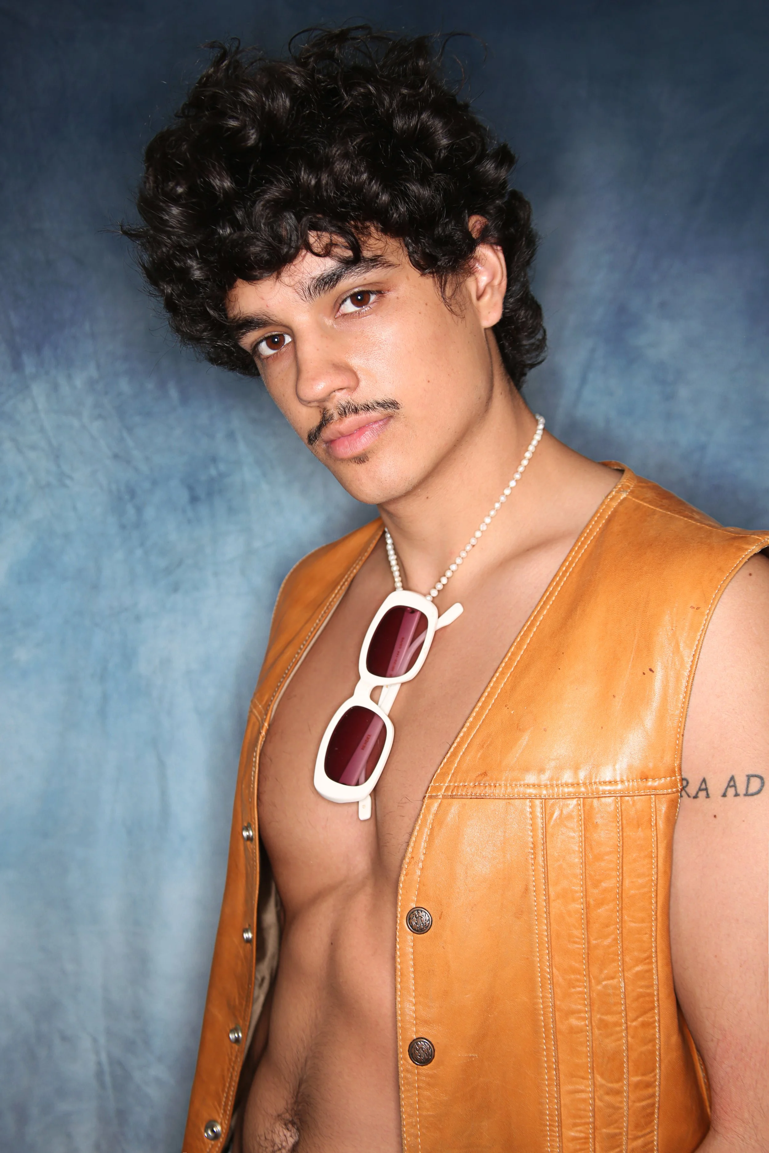A young man with curly dark hair and light skin, wearing a tan leather sleeveless vest, sunglasses hanging from his neck, and a beaded necklace, looking at the camera against a dark blue mottled background.