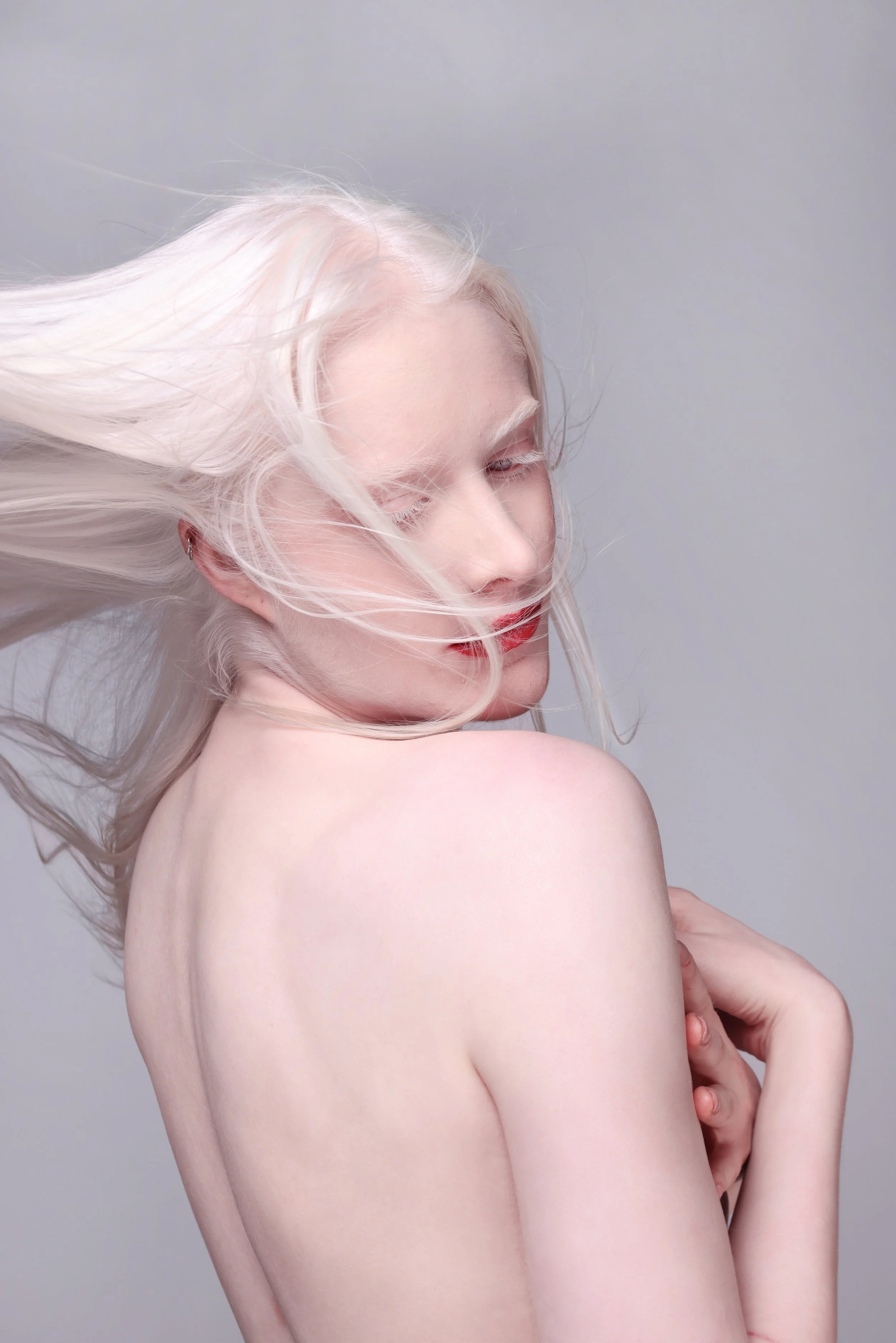 A nude woman with long platinum blonde hair blowing to the left, with her eyes closed, standing against a plain grey background.