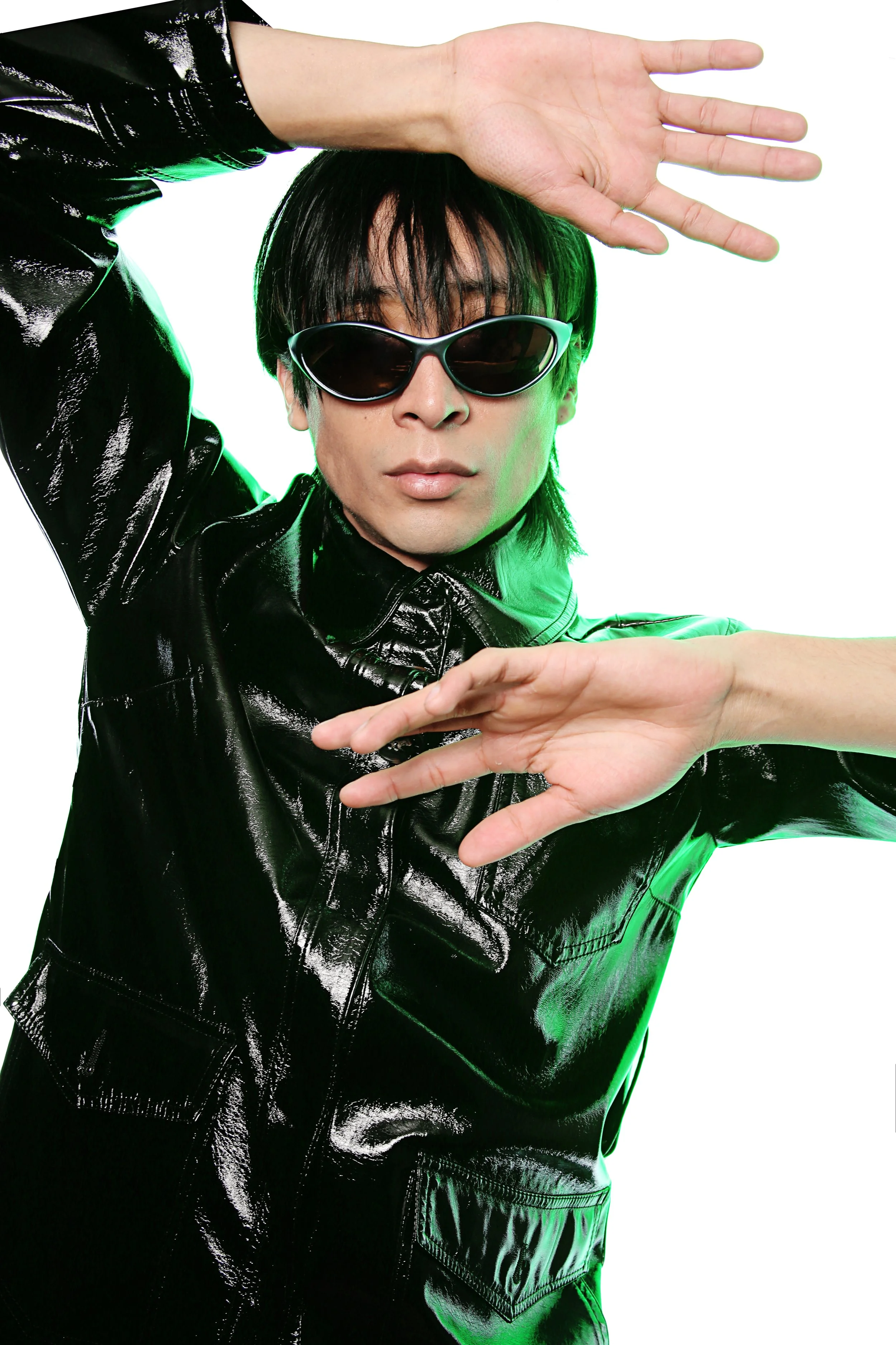 A man with dark hair and light skin wearing dark sunglasses and a shiny black leather jacket, posing with one arm raised and the other extended horizontally.