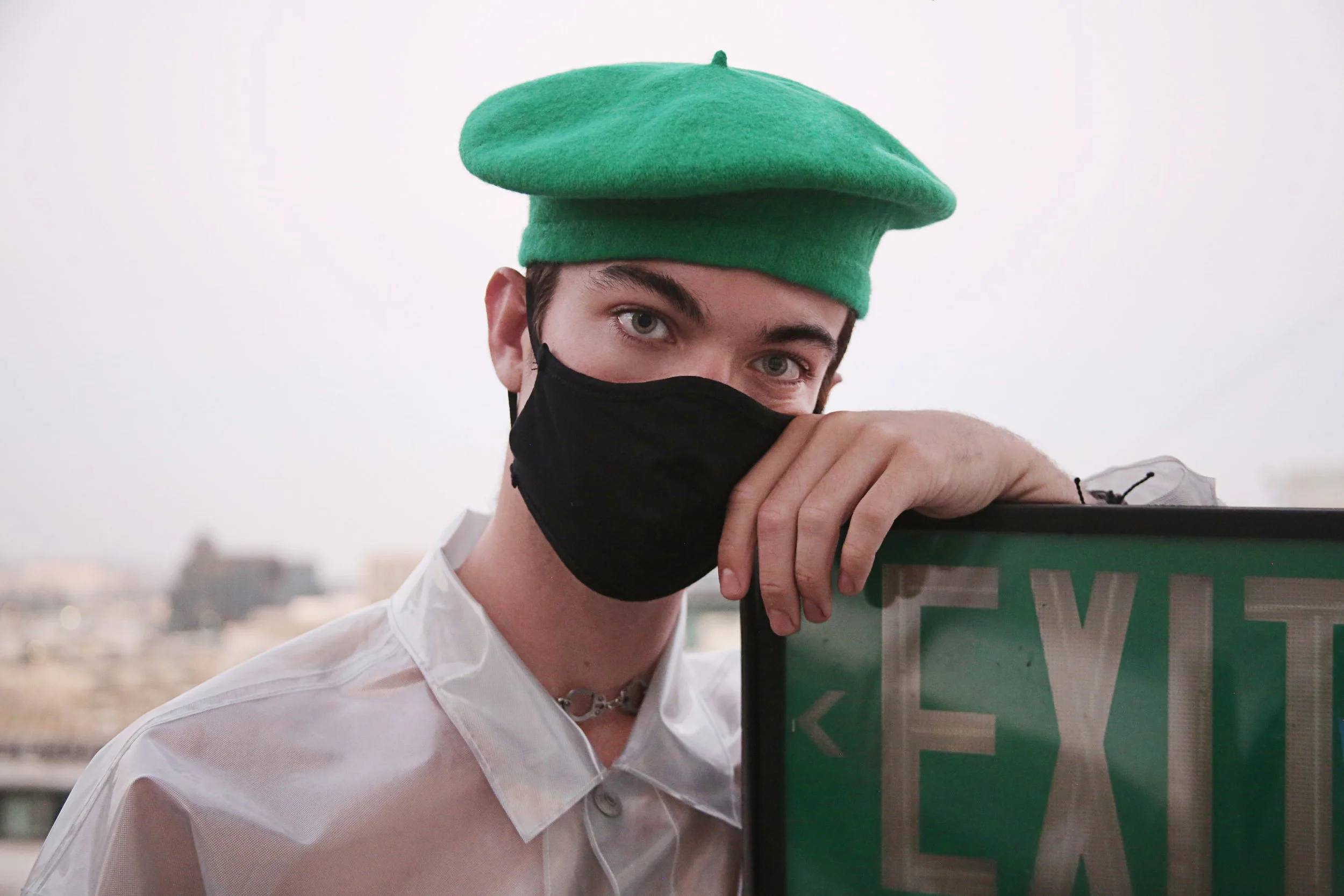 A person with green eyes wearing a green beret, black face mask, and white shirt, leaning on a green sign outdoors with an urban background.