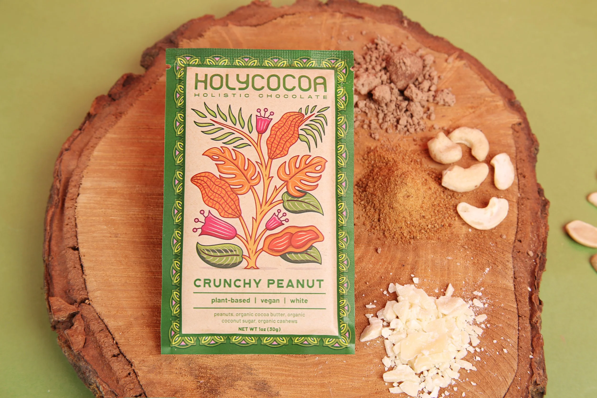 A chocolate treat with a terracotta color, topped with crushed white nuts and a spice-like powder, placed on a wooden surface. Partly covered by a green and white box labeled "HolyCoca Holistic Chocolate" with colorful botanical illustrations.