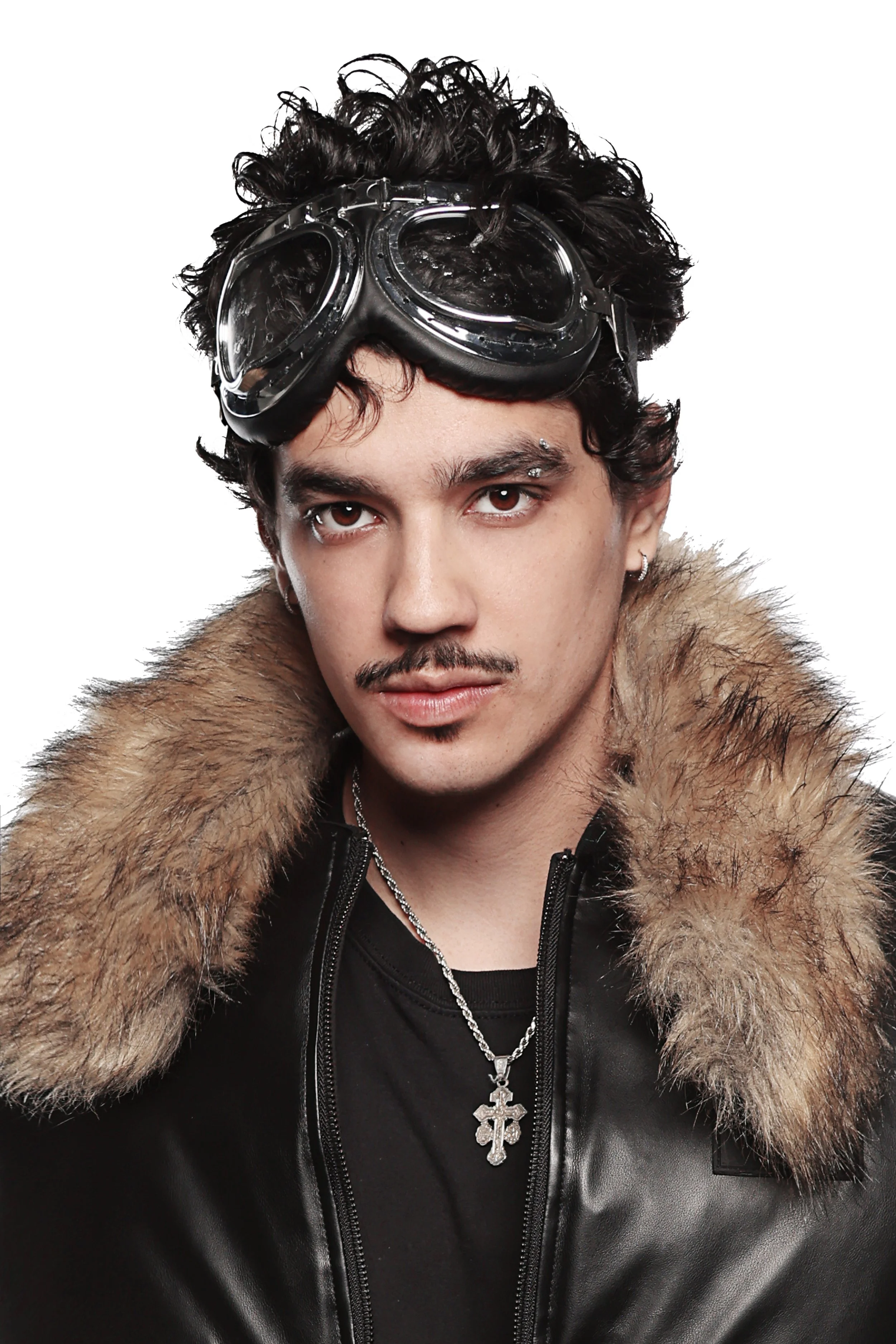 Young man with dark curly hair wearing goggles, jewelry, a fur-lined black leather jacket, and a large cross necklace, with a serious expression.
