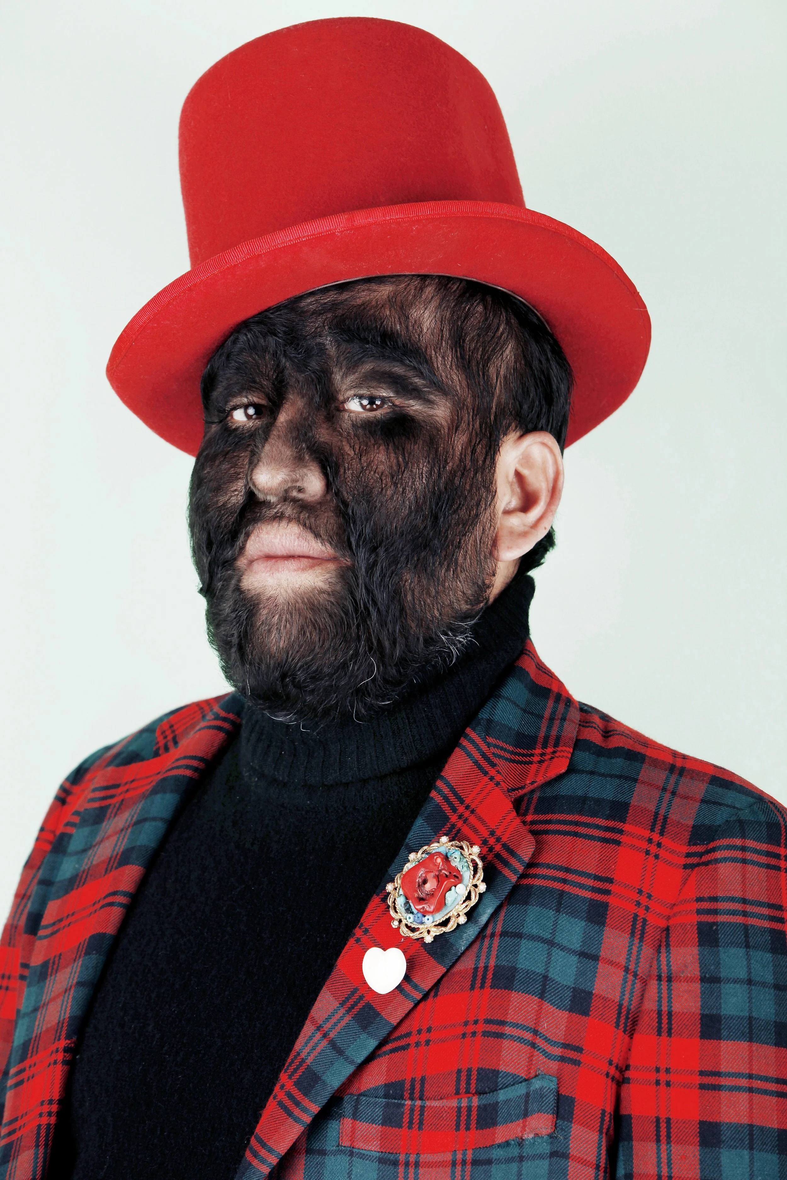 A person with dark hair and beard wearing a red plaid jacket, a black turtleneck, a red top hat, and a decorative brooch on the jacket, against a light background.