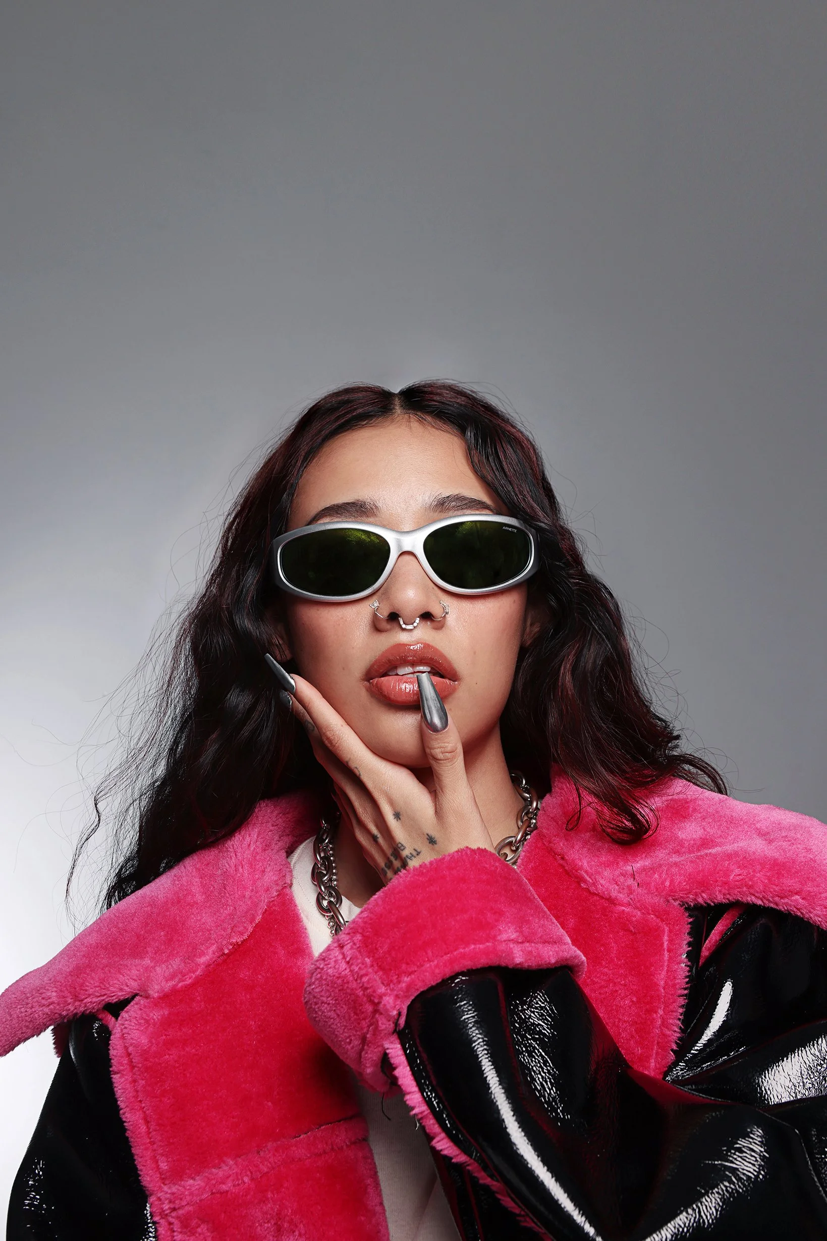 A woman with dark wavy hair, wearing white sunglasses, a pink fuzzy jacket, and a silver chain necklace, poses with her hand touching her chin against a gray background.