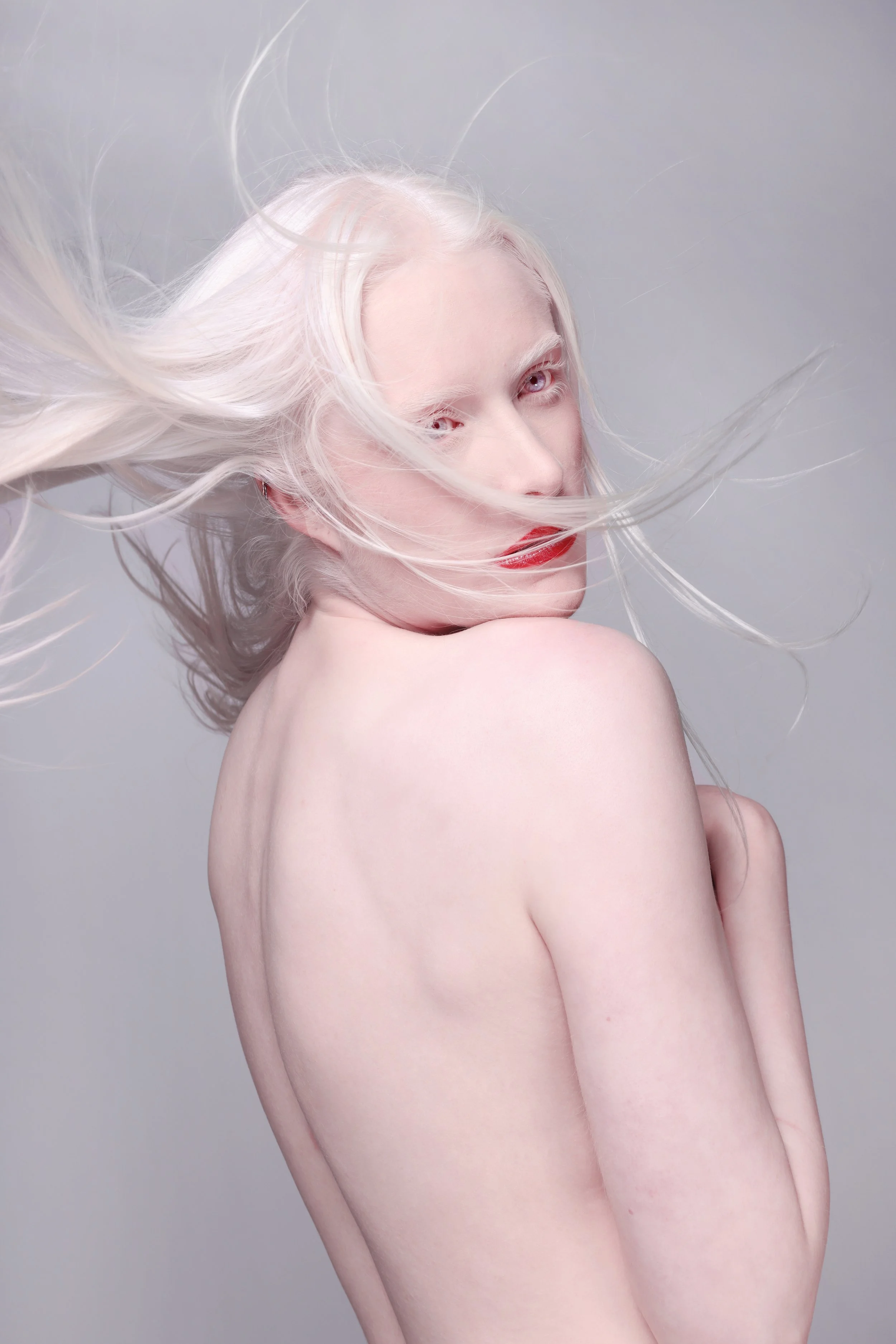 A woman with platinum blonde hair and red lipstick, posing with her back and shoulder exposed, hair flowing around her face against a light gray background.