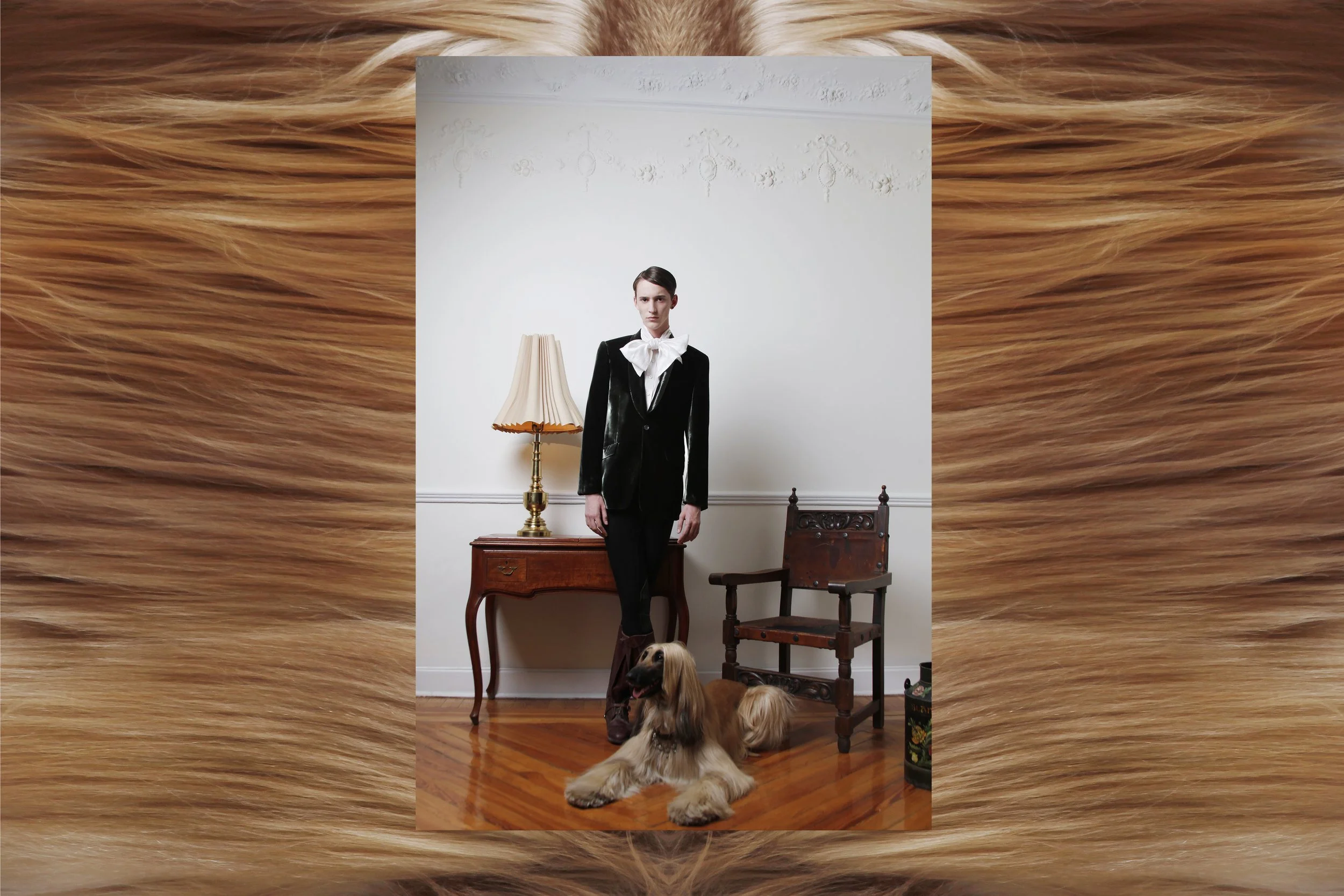 A woman dressed in black and white standing in a room with vintage furniture, including a wooden table, a chair, and a lamp, with a Cavalier King Charles Spaniel lying on the wooden floor, and a portrait of a dog on a metal container.