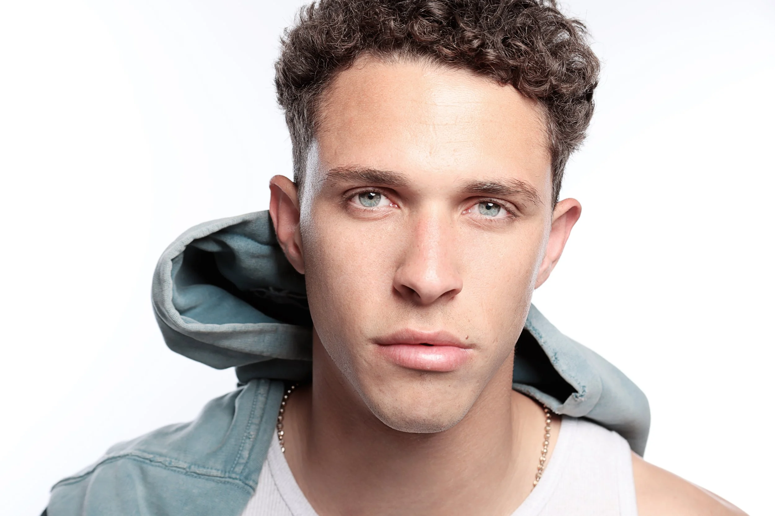 A young man with short curly brown hair and blue eyes wearing a white tank top, a gray hoodie, and a chain necklace, looking at the camera with a neutral expression against a white background.