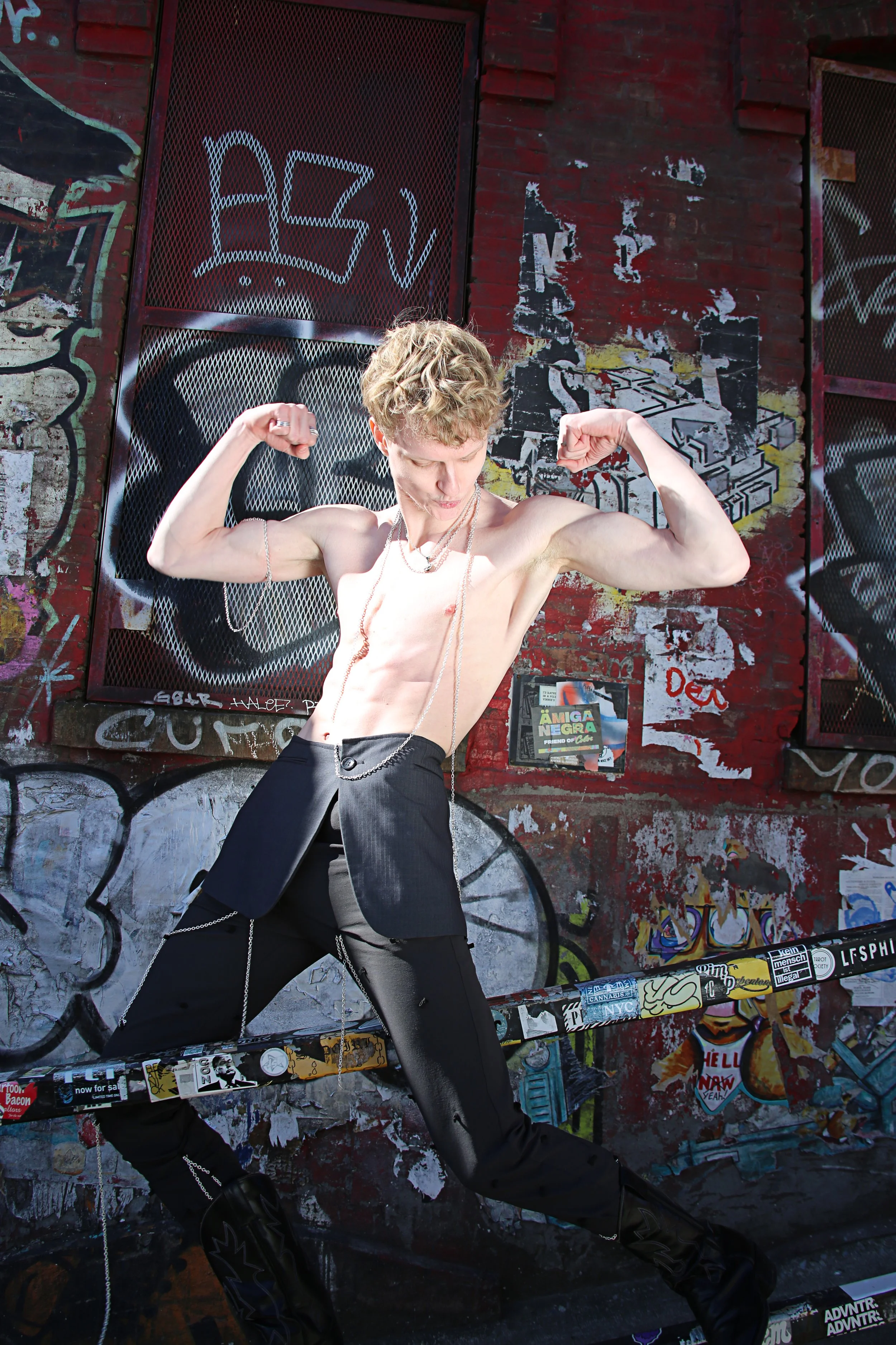 A shirtless person with short, curly blond hair flexes their arms while standing in front of a graffiti-covered brick wall. They wear black pants with chains and jewelry accessories, including a necklace and rings.