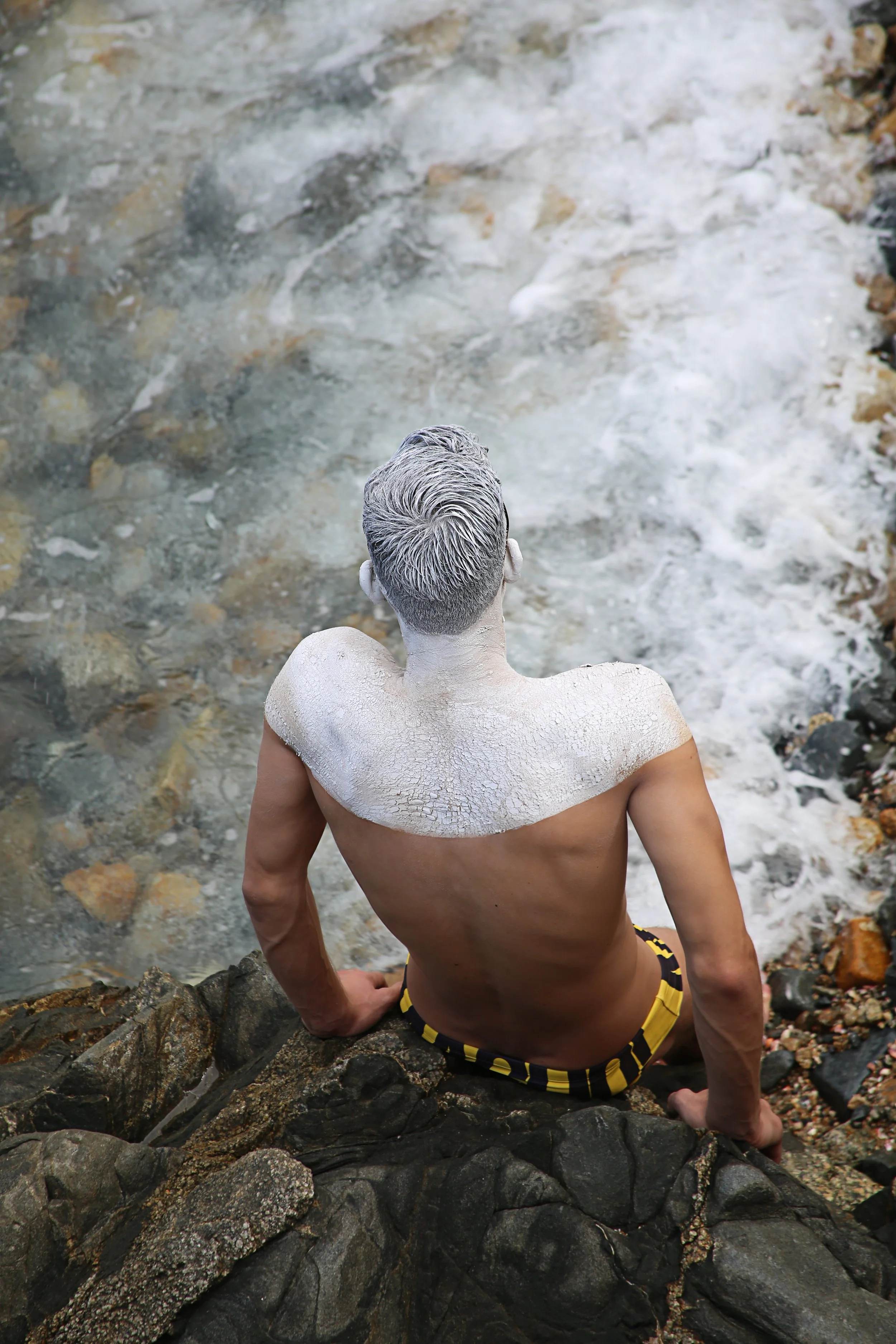 A man with gray hair and tanned skin, wearing yellow and black swim trunks, is sitting on black rocks near the ocean, facing away from the camera with water and foam behind him.