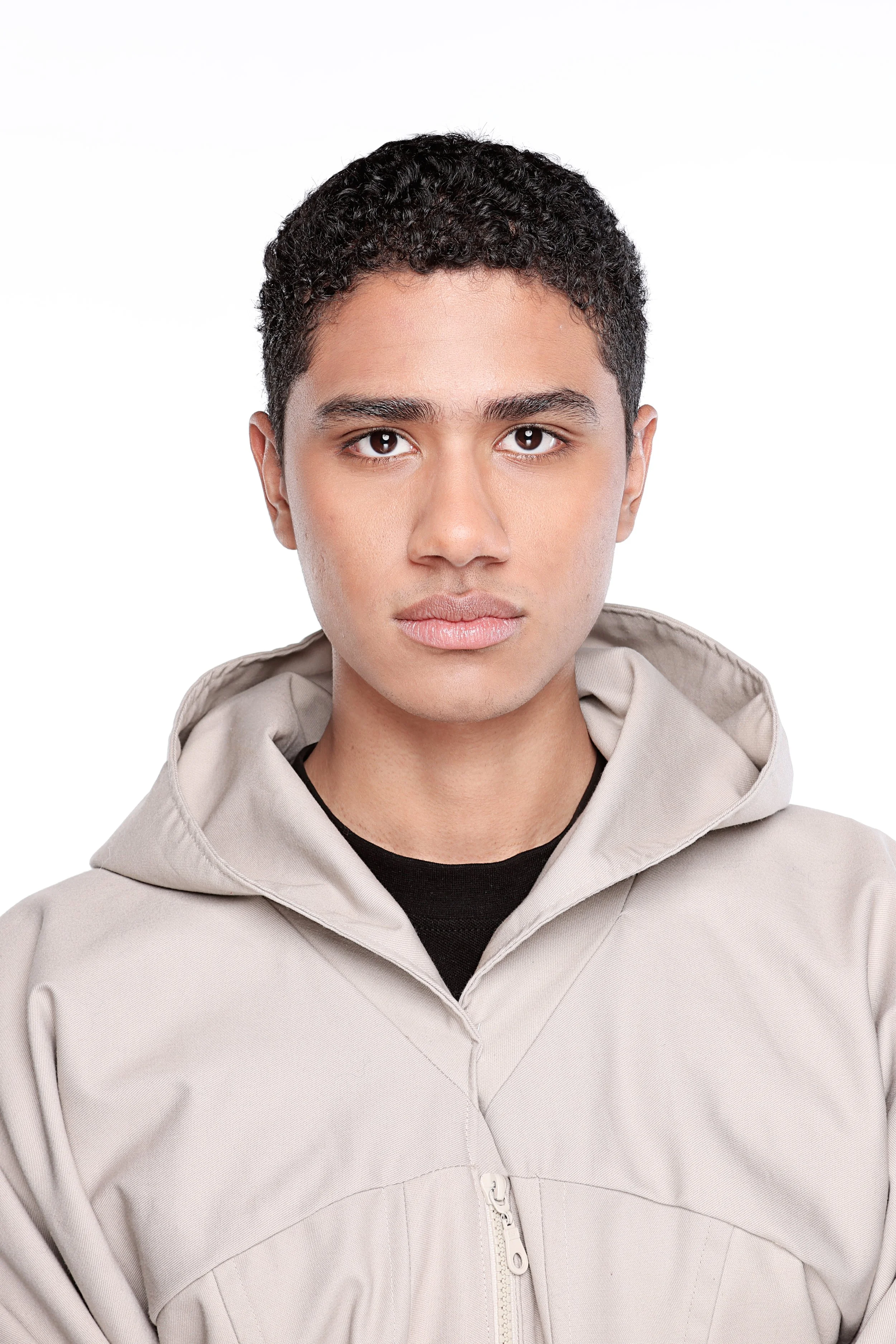 A young man with short curly black hair and tan skin wearing a beige jacket with a black shirt underneath, standing against a plain white background.