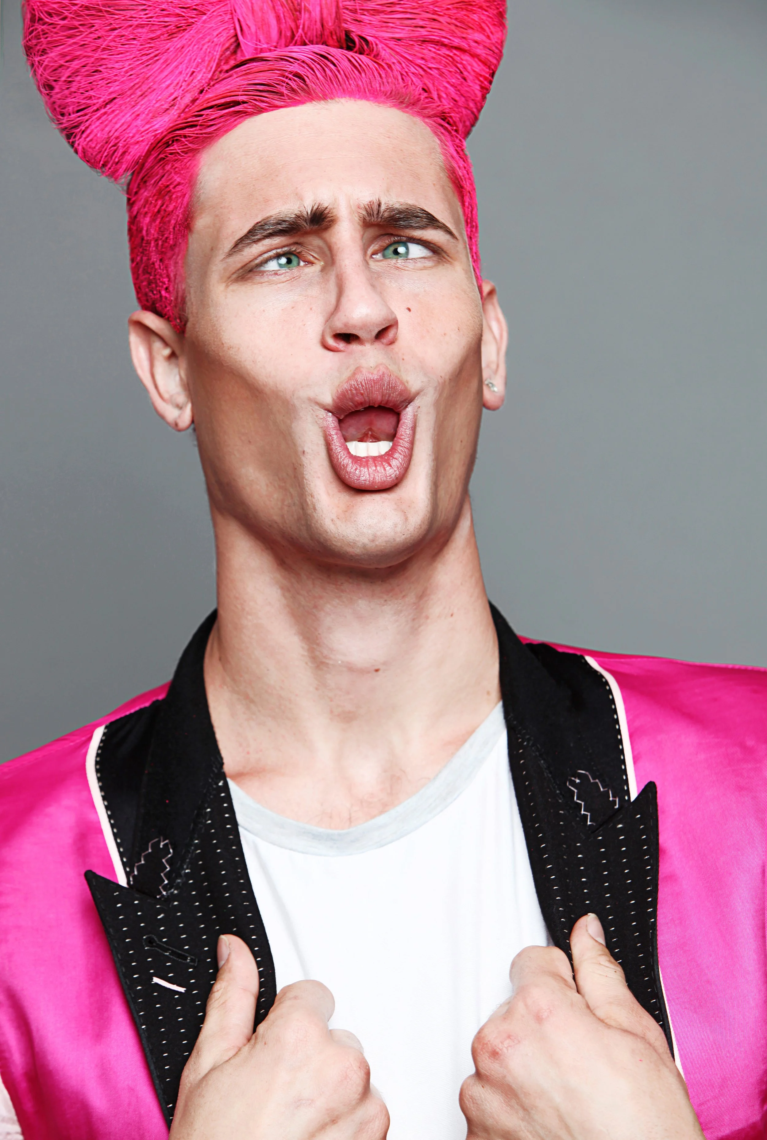 Person with bright pink hair styled in an elaborate updo, making a surprised or excited facial expression with lips pursed and eyes wide open, wearing a black collared shirt with white dots and a pink jacket, holding the lapels with both hands.