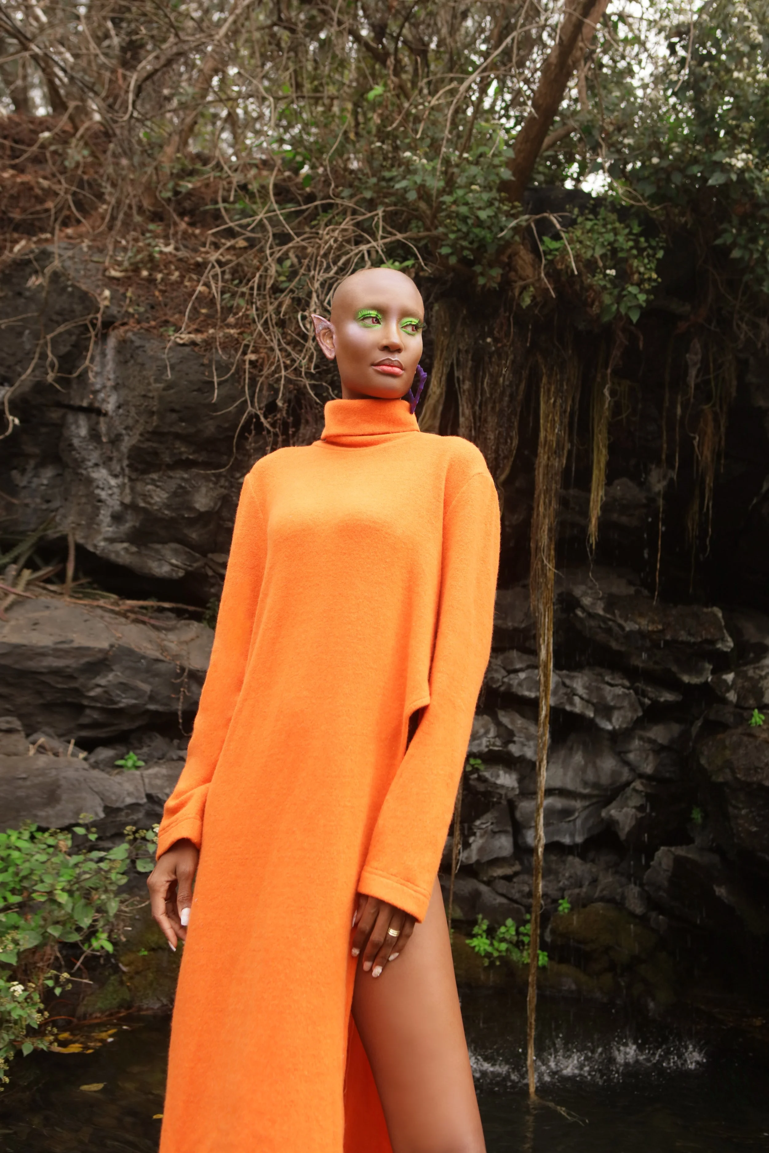 A woman with a bald head and elf-like ears wearing an orange garment standing outdoors near a rocky water feature and dense vegetation.