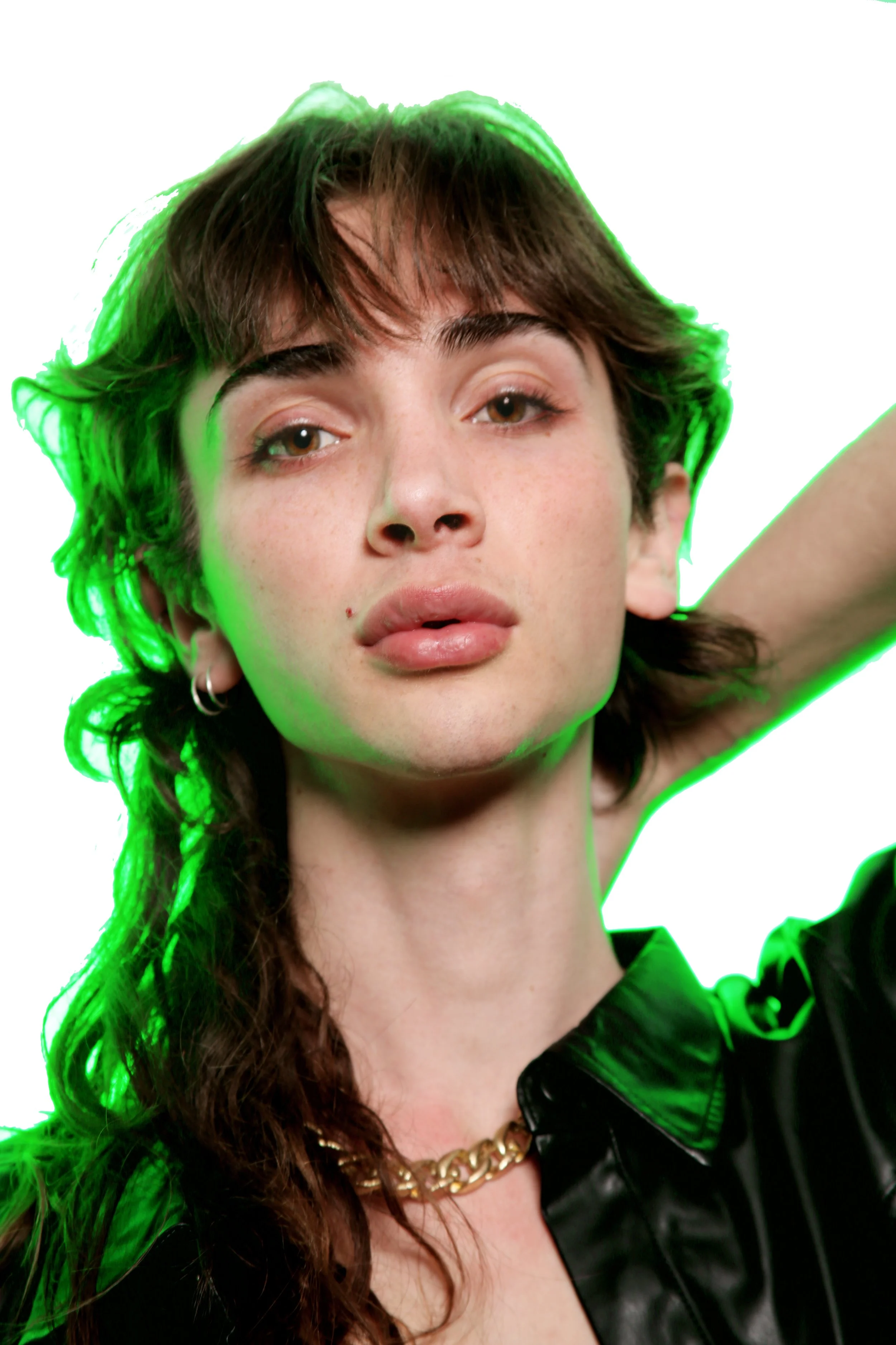 A woman with brown hair, styled in loose waves, wearing a black leather jacket and a gold chain necklace, with a white background and a green glow outlining her hair.