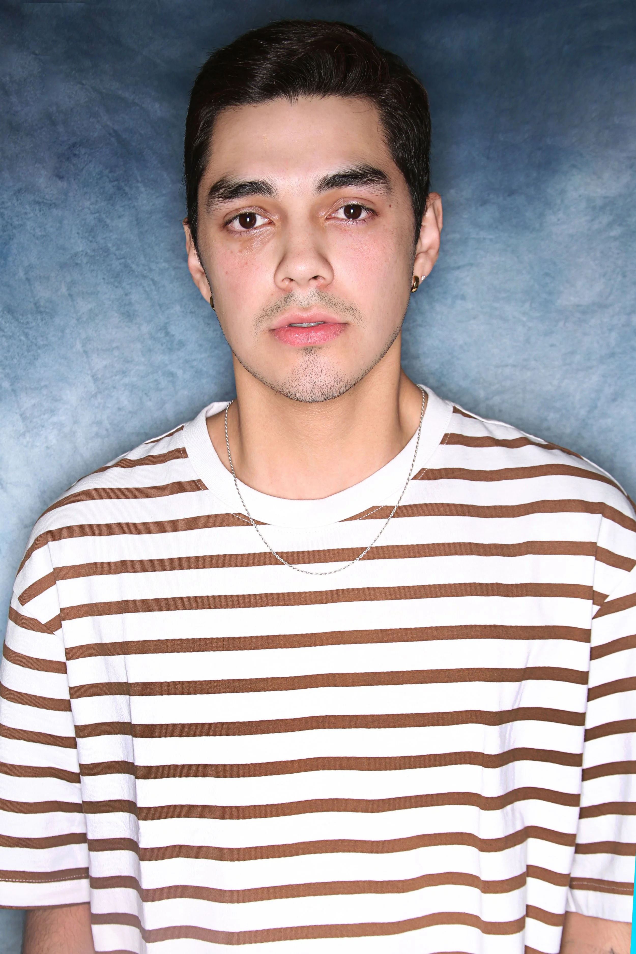 Close-up of a young man with short dark hair, wearing a white and brown striped T-shirt, standing against a blue textured background.