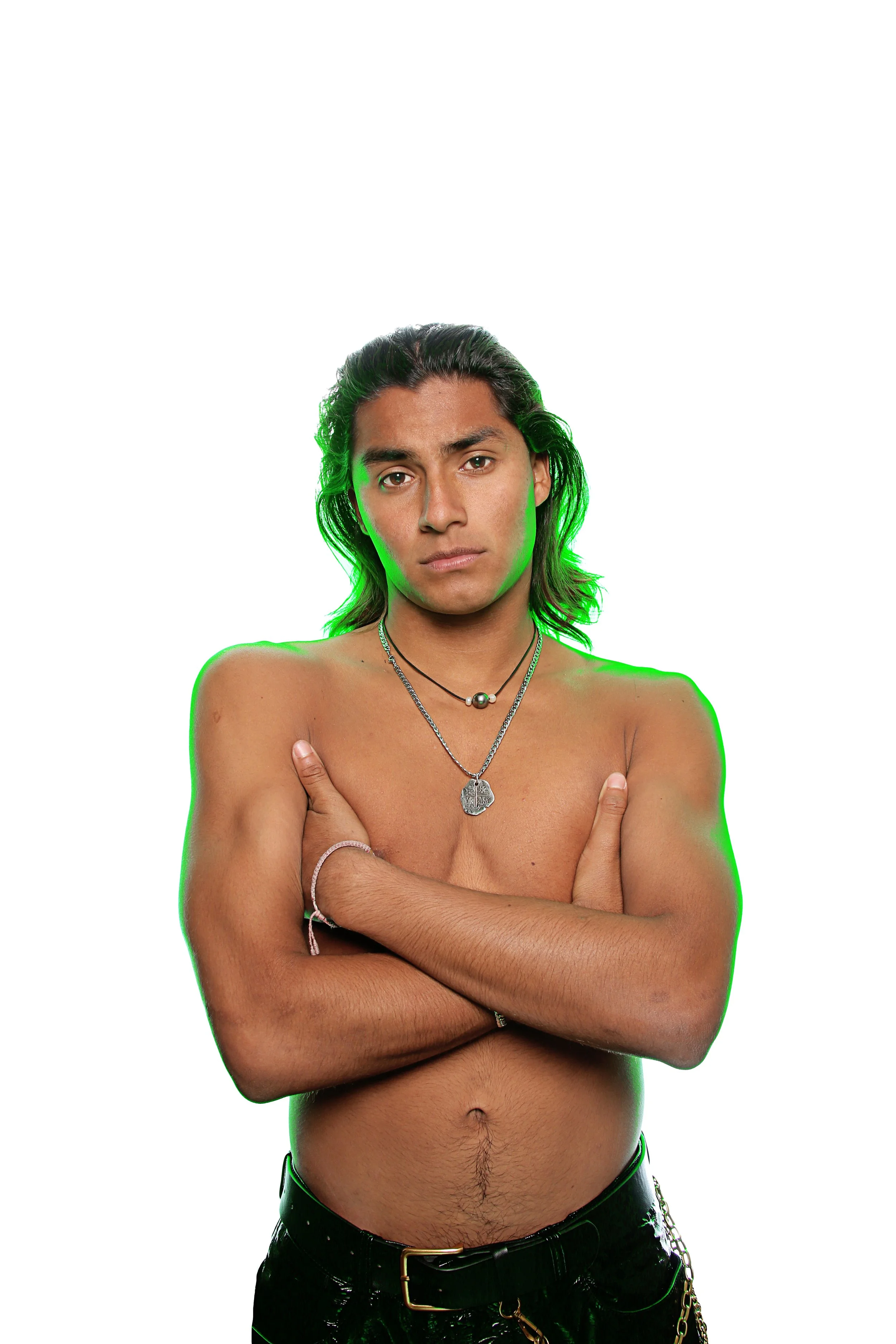 A young man with long dark hair and a serious expression, standing shirtless against a white background, with his arms crossed over his chest