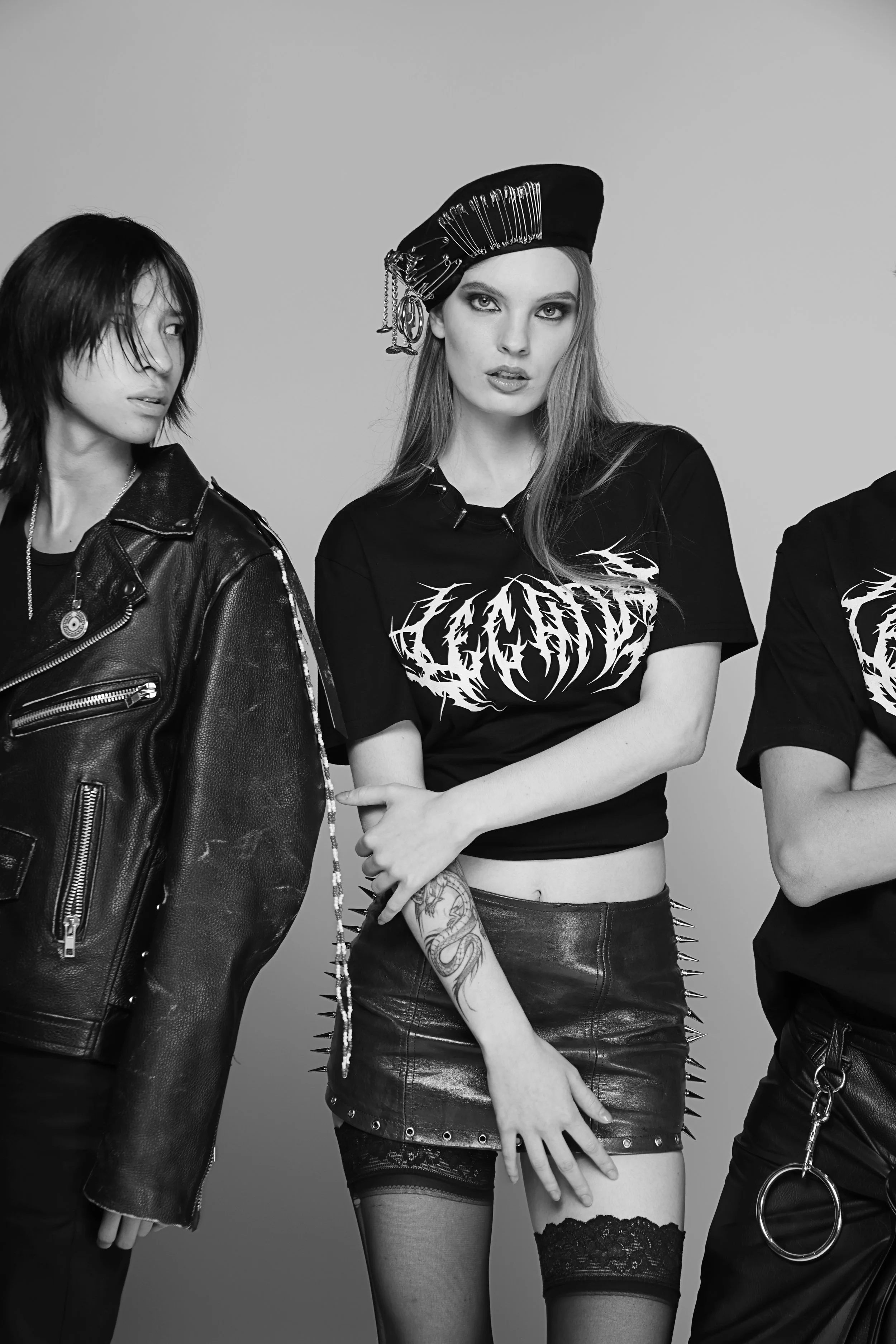 Black and white photo of three people dressed in punk or goth style clothing, with the focus on the middle woman who has long hair, a studded leather skirt, lace top, and striped thigh-high stockings. The woman wears a hat with chains and safety pins