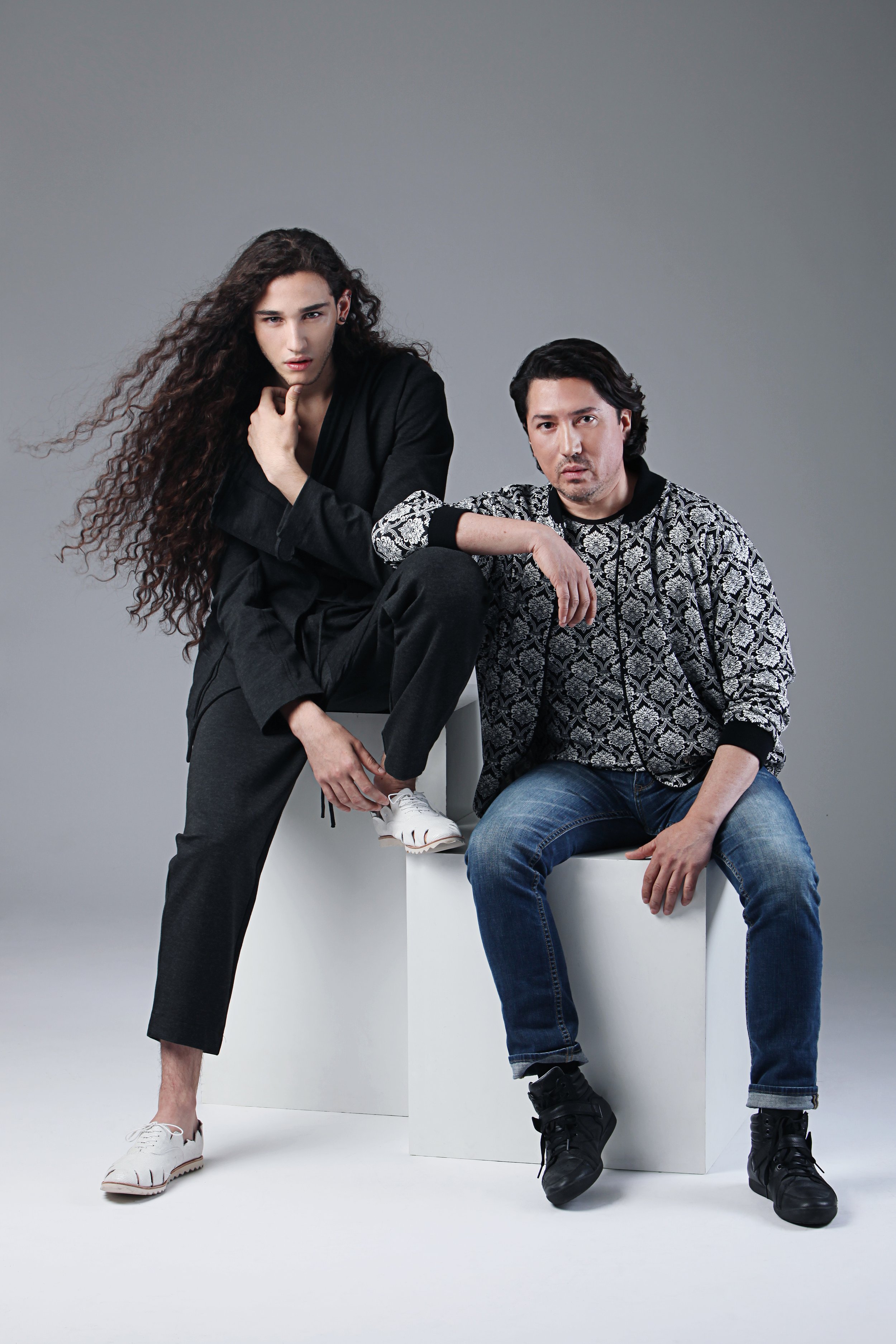 A man and a woman posing together against a gray background, with the woman standing and the man sitting on a white block.
