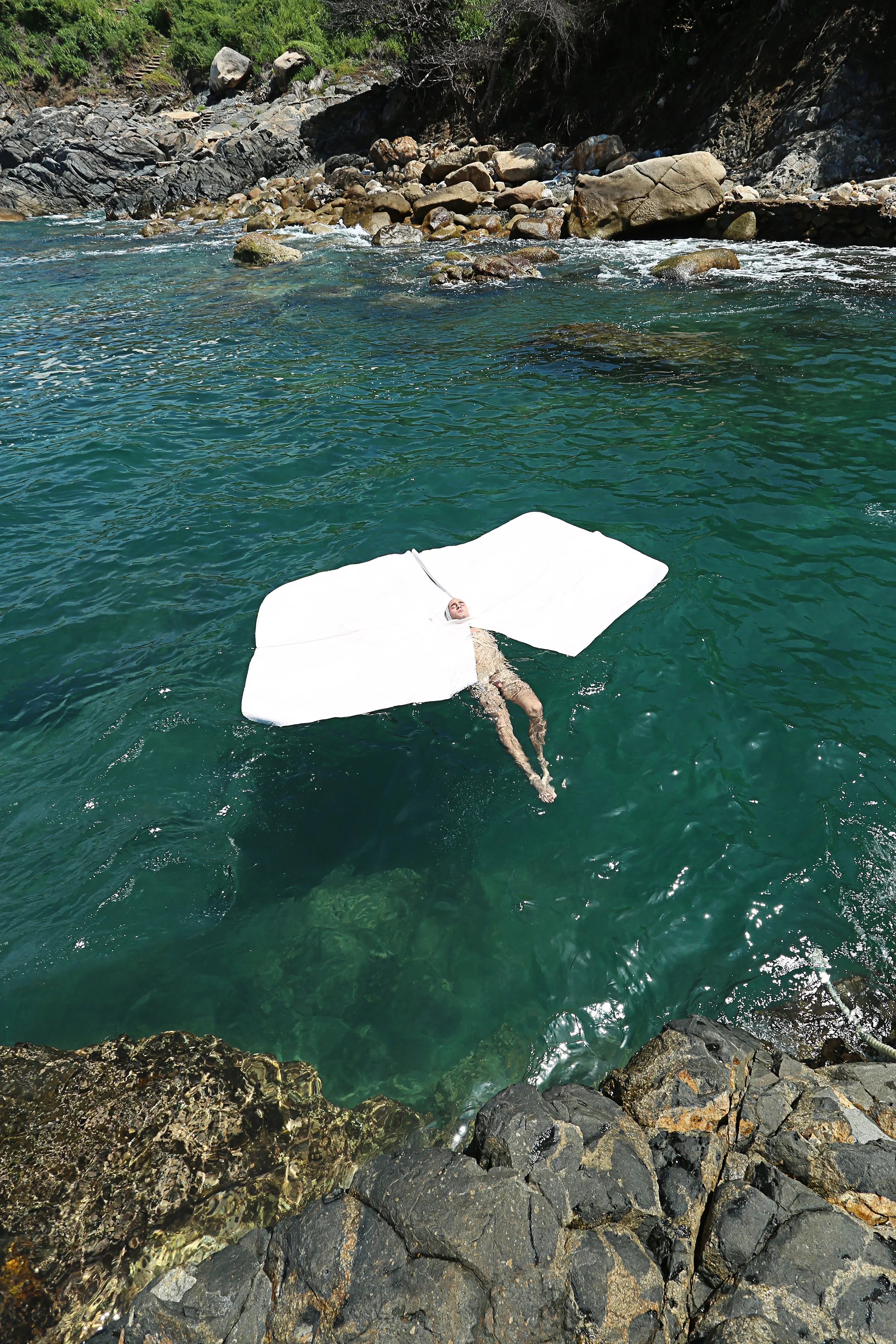 A person floating on their back in a clear river, held by a white floating device, with rocks and greenery along the riverbank.