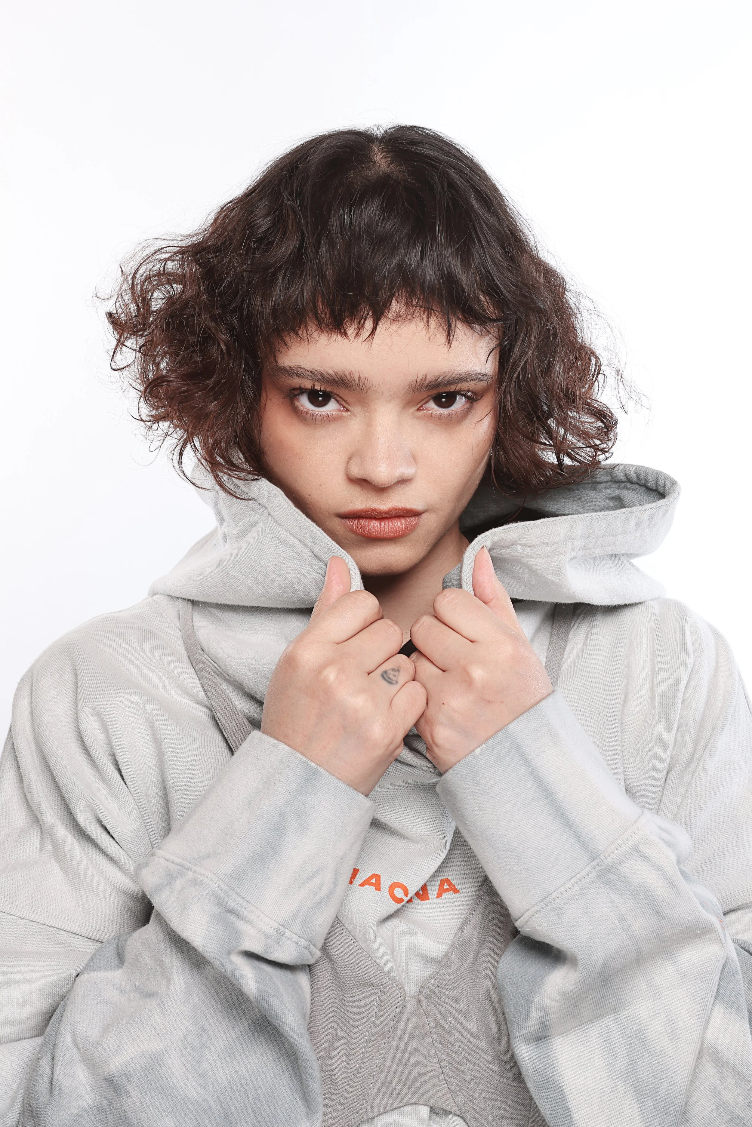 A young woman with short, curly dark hair, wearing a light gray hoodie, looking directly at the camera with a serious expression. She is holding the edges of her hoodie near her face with both hands.