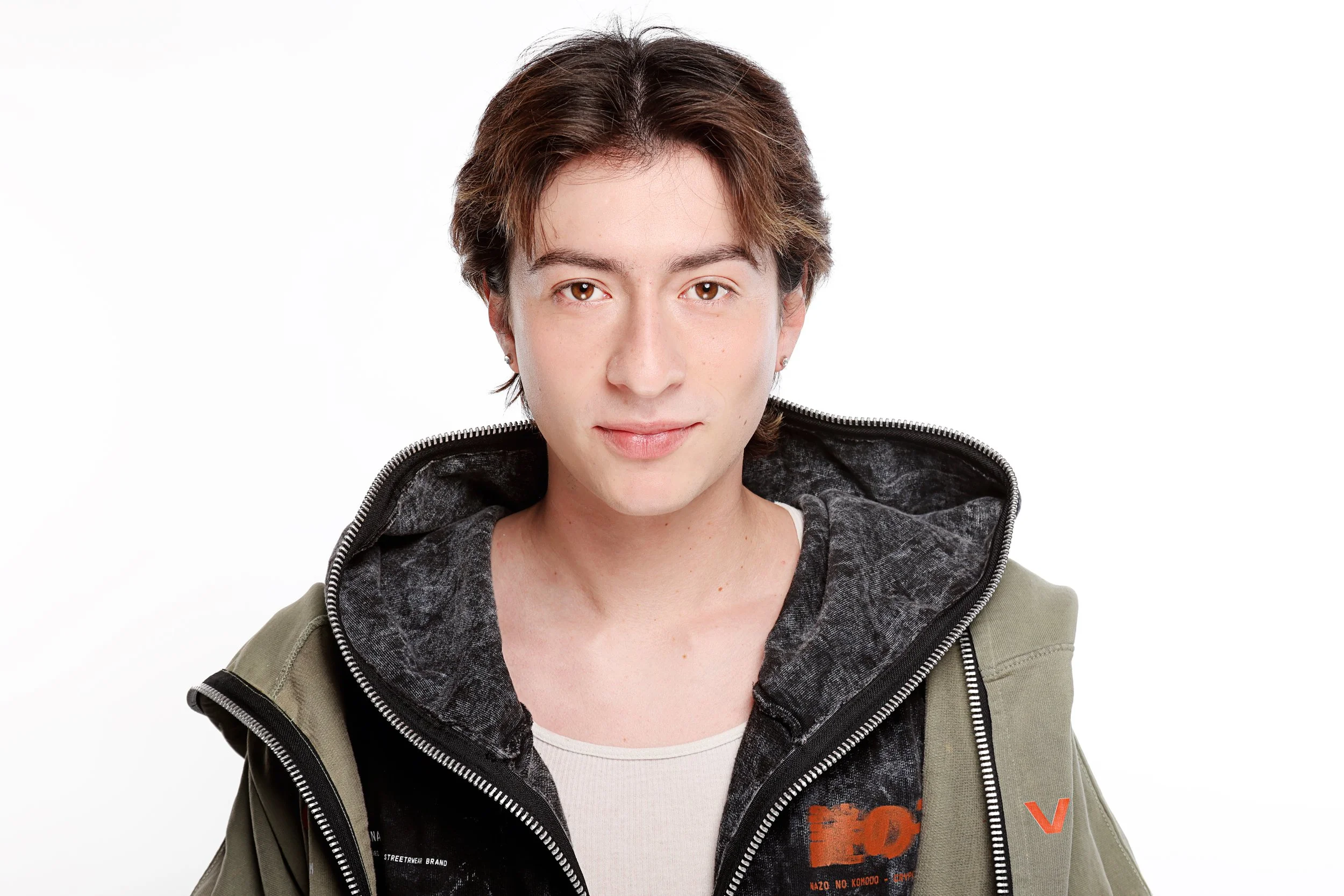 A young person with short brown hair and light skin, wearing a layered outfit including a gray and black hoodie with a zipper and a tan jacket, against a white background.