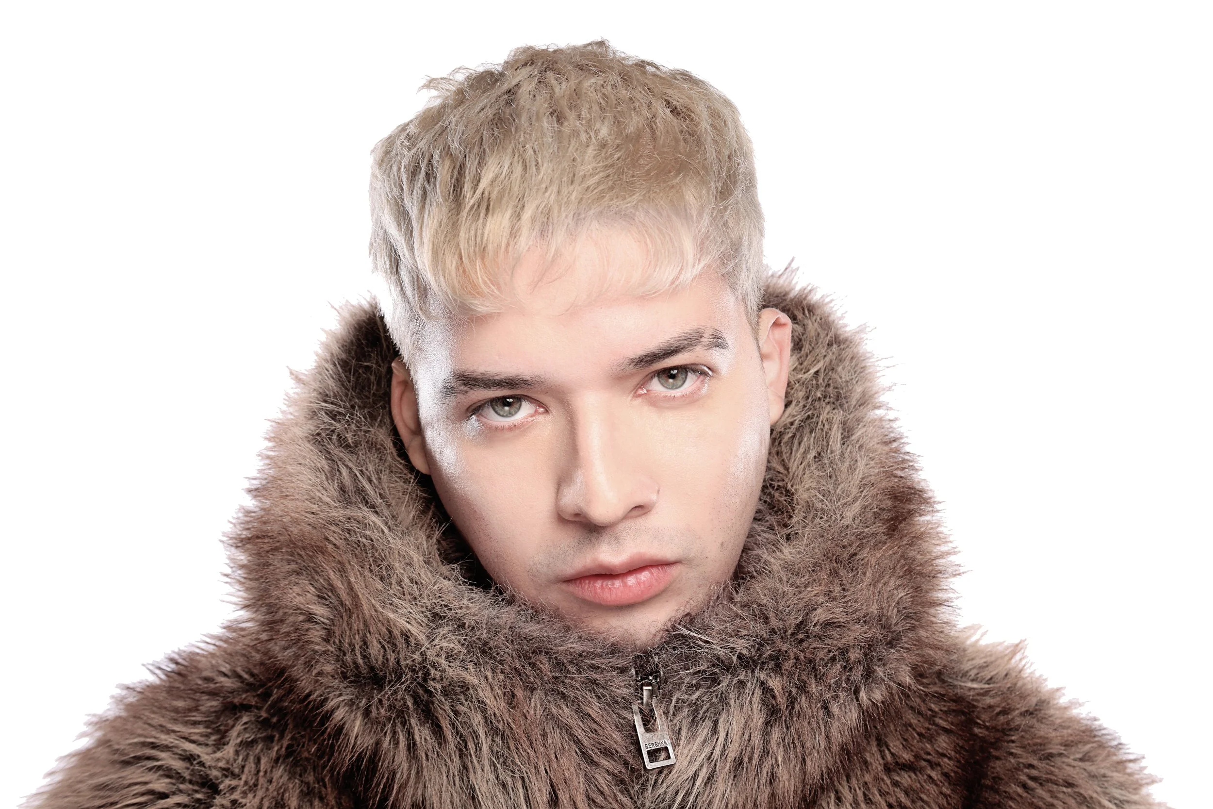Close-up of a person wearing a brown fur coat with a zipper, with short, blonde hair and light eyes, against a white background.