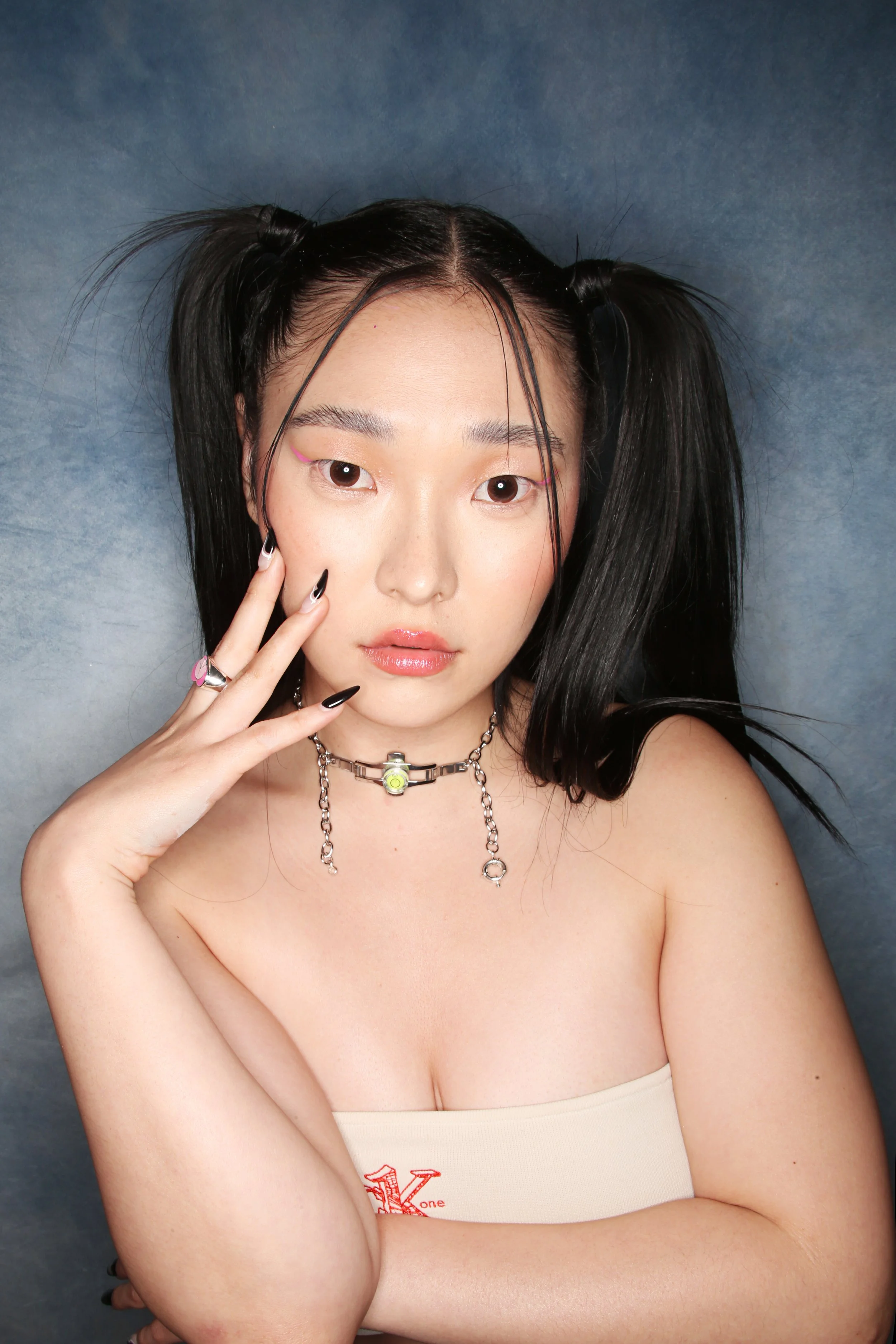 A young woman with black hair styled in pigtails, wearing a beige tube top, and posing with her hand near her face against a dark blue background.