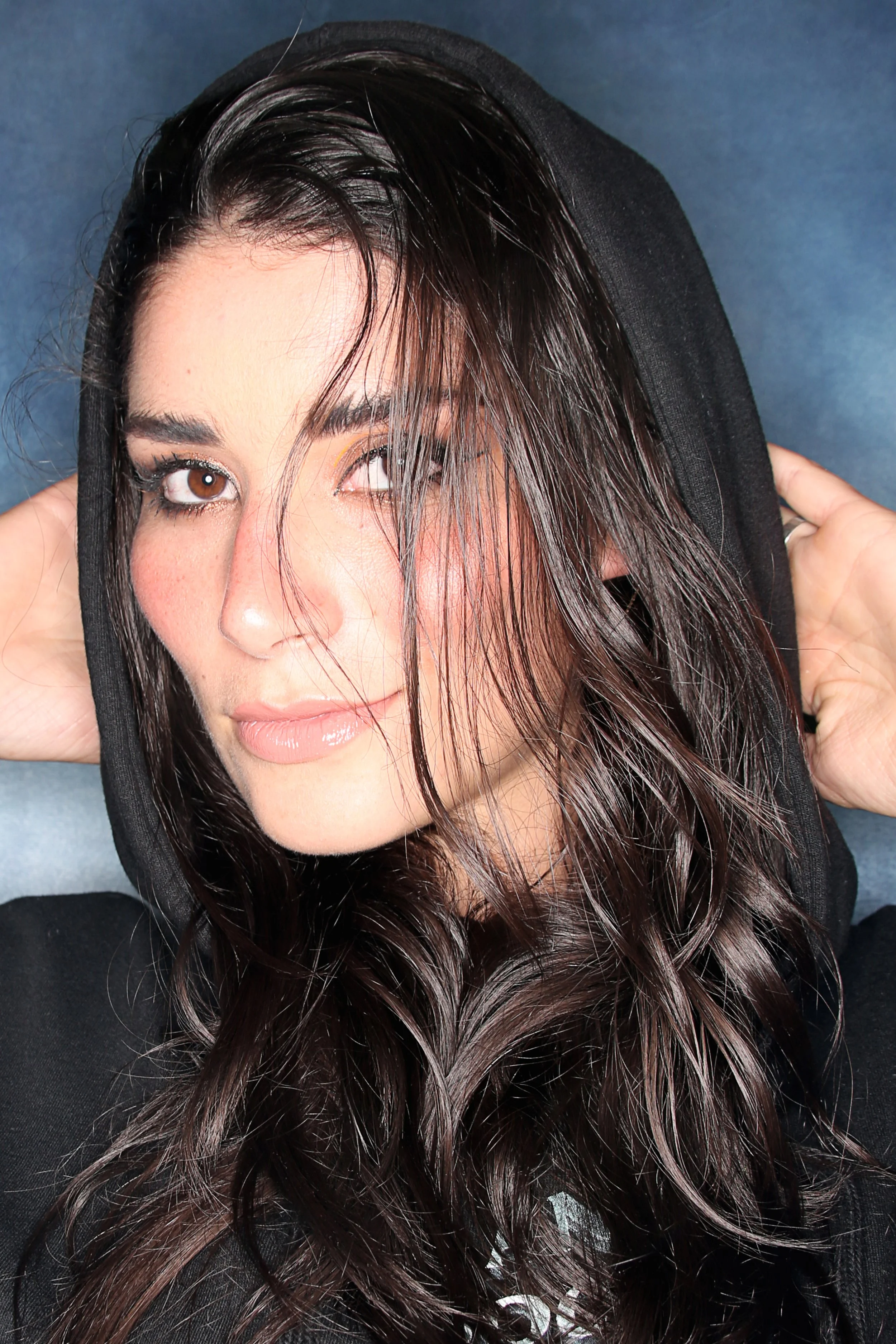 A woman with wet dark hair wearing a black hoodie with the hood up, looking at the camera. She is touching the hood with both hands, and the background is a plain blue surface.
