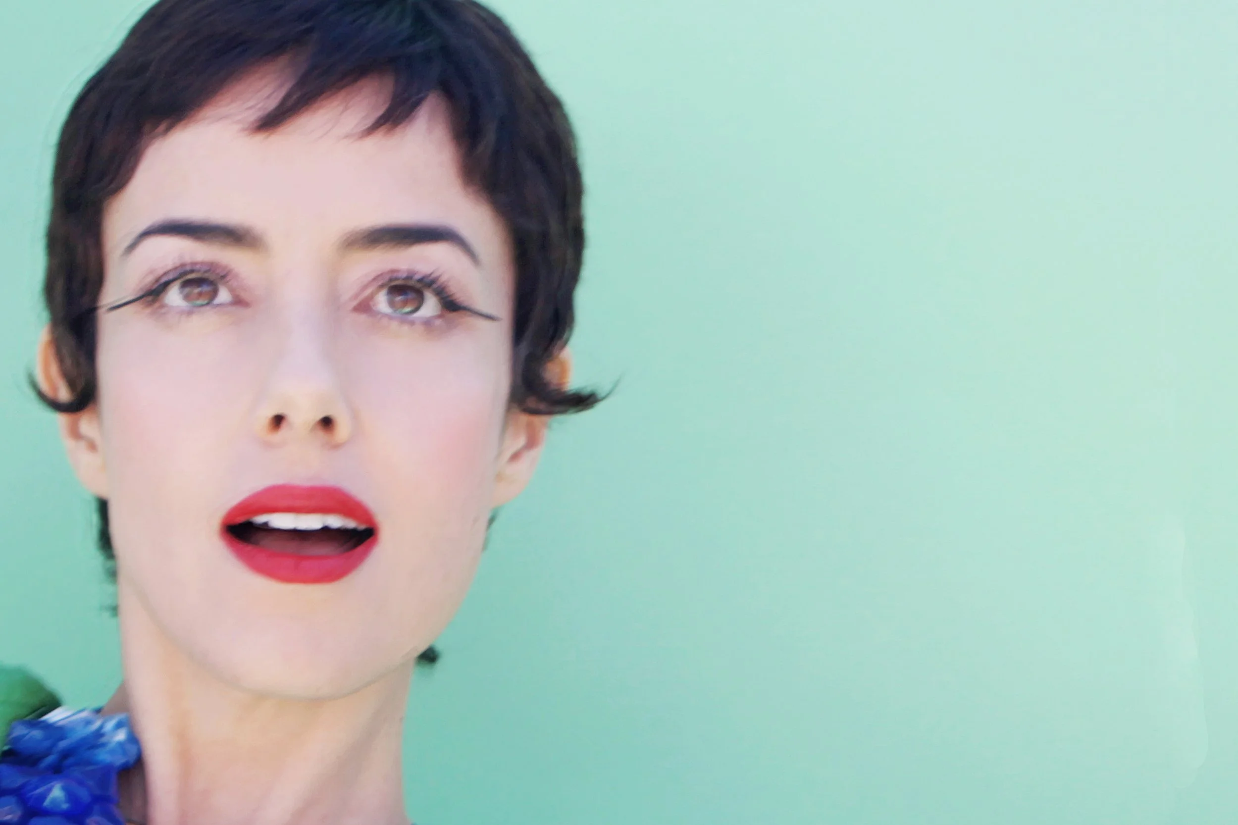 Close-up of a woman with short dark hair, red lipstick, and open mouth, against a light green background.