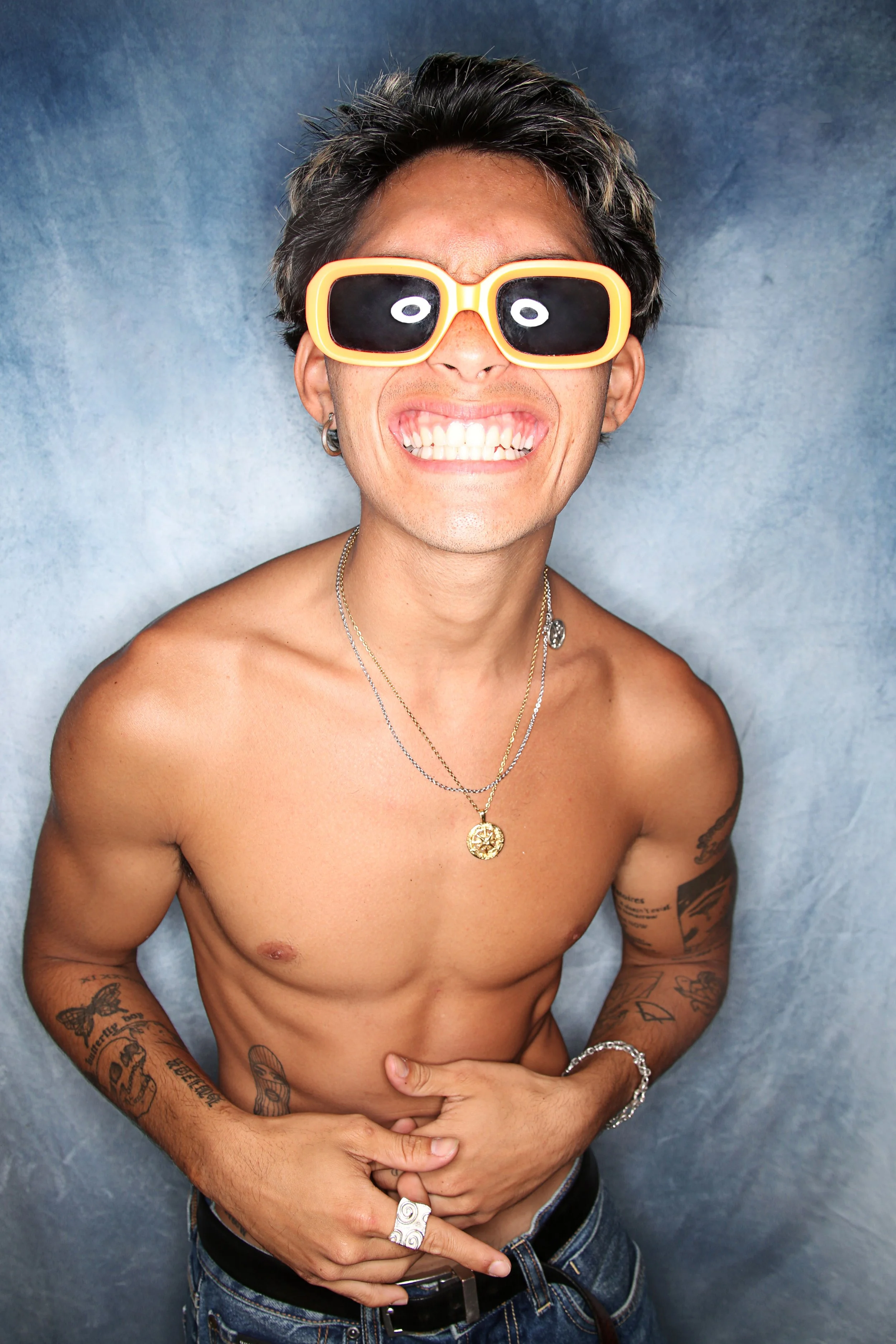 A shirtless man with dark hair and tattoos, wearing sunglasses with a cartoonish style, smiling broadly, and holding his stomach. He is accessorized with jewelry including necklaces, rings, earrings, and bracelets, and standing in front of a grayish-