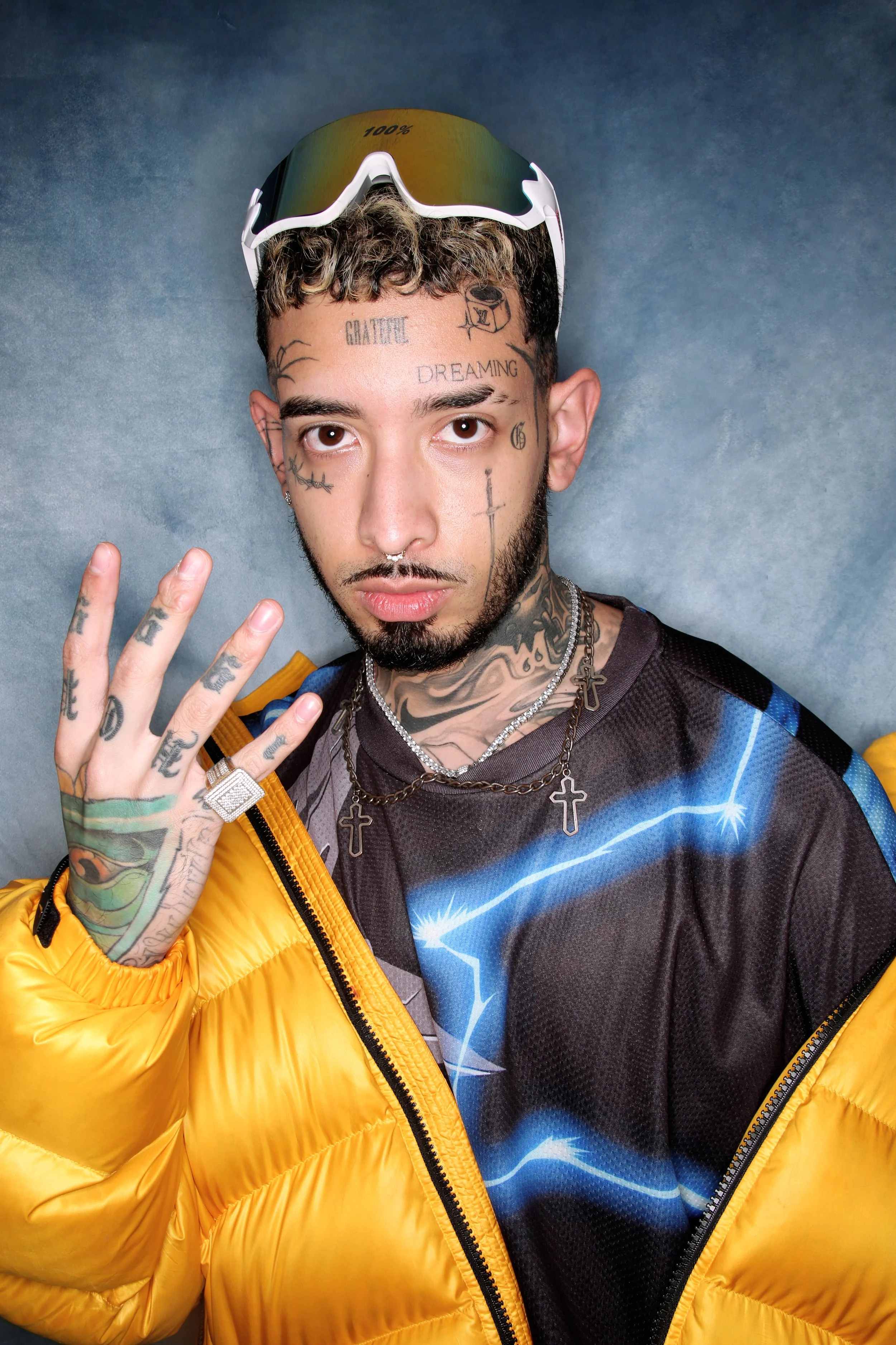 A young man with tattoos on face, neck, and hand, wearing a yellow puffer jacket, colorful sports shirt, multiple necklaces, and sunglasses on his head, making a peace sign.