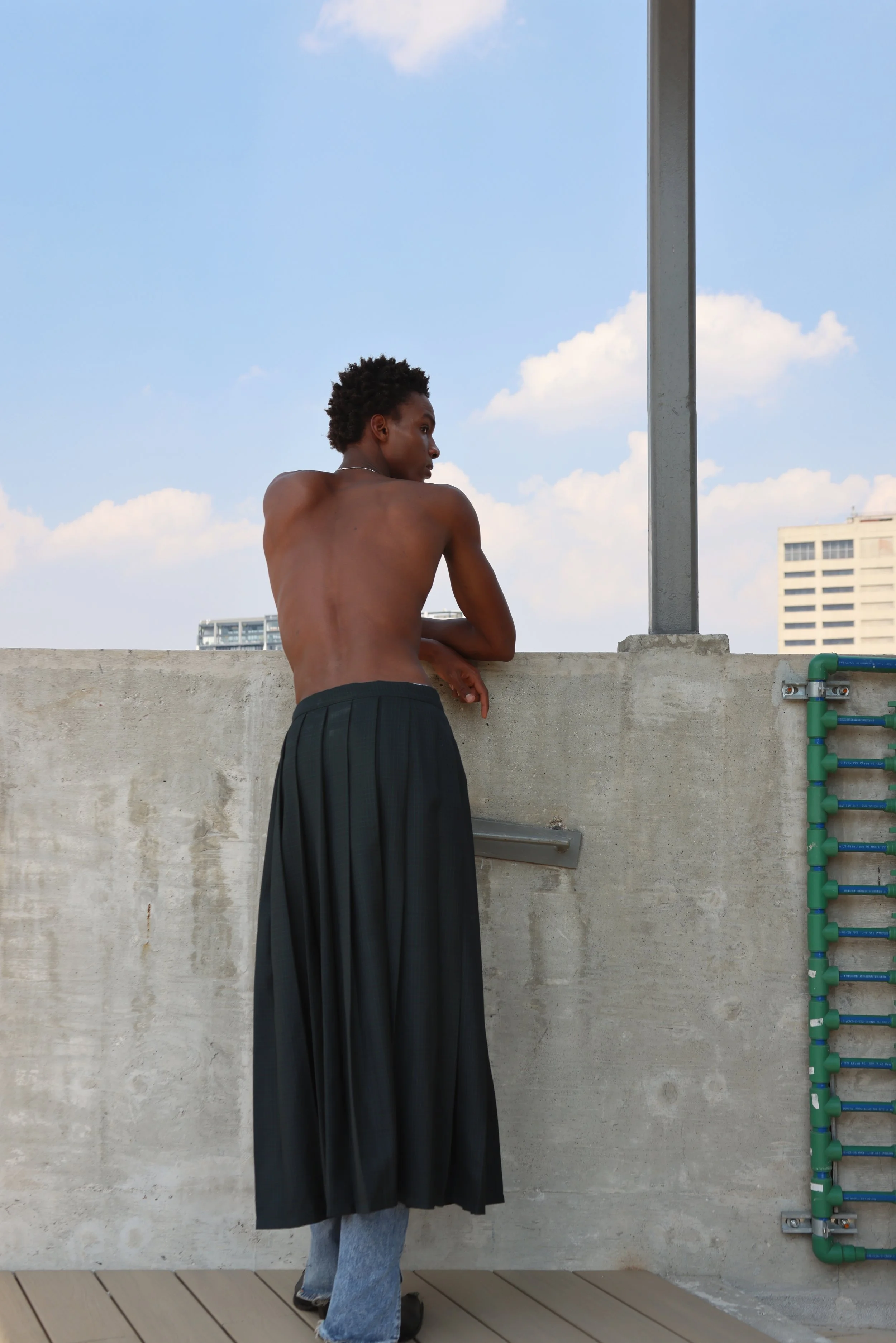 A person with short curly hair and dark skin stands on a rooftop, leaning on a concrete wall, looking to the side. They are wearing black pants and gray shoes, no shirt. The sky is partly cloudy, and a tall building is visible in the background. Ther
