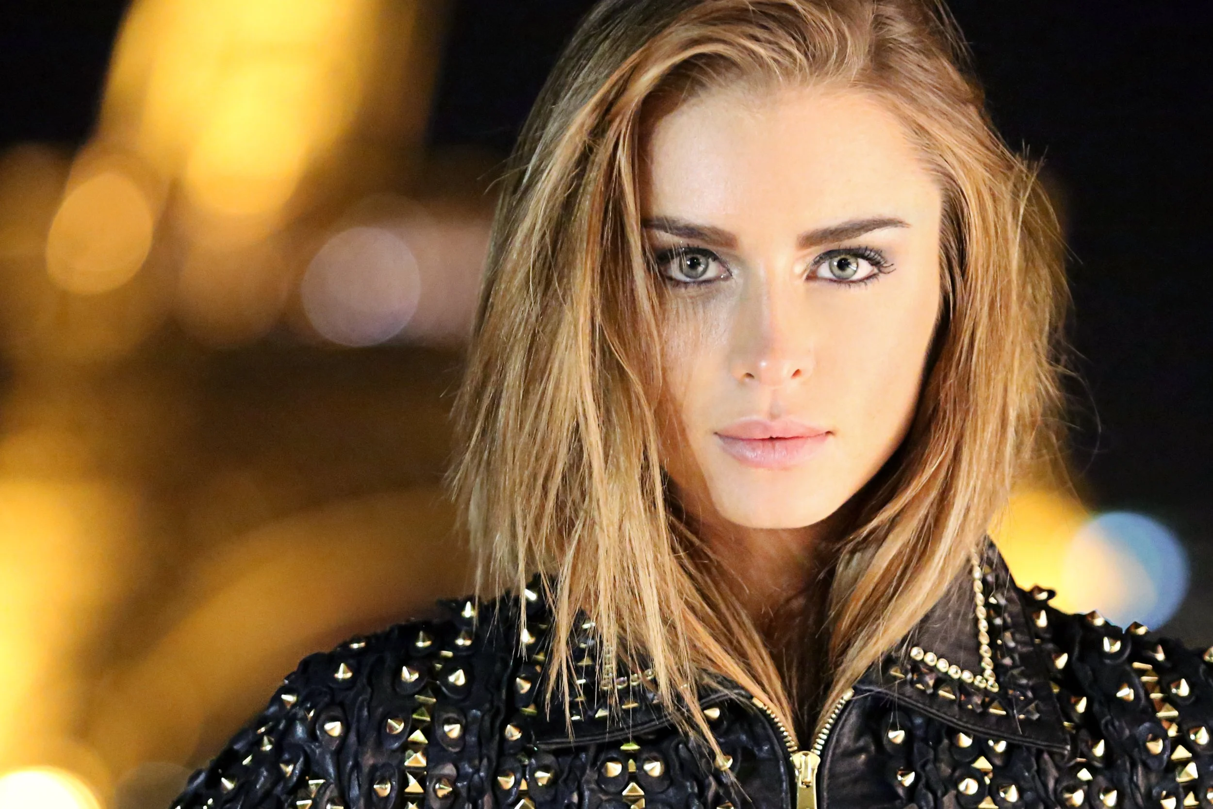 Close-up of a young woman with long, wavy, reddish-blonde hair and light-colored eyes, wearing a black leather jacket with silver studs, outdoors at night.