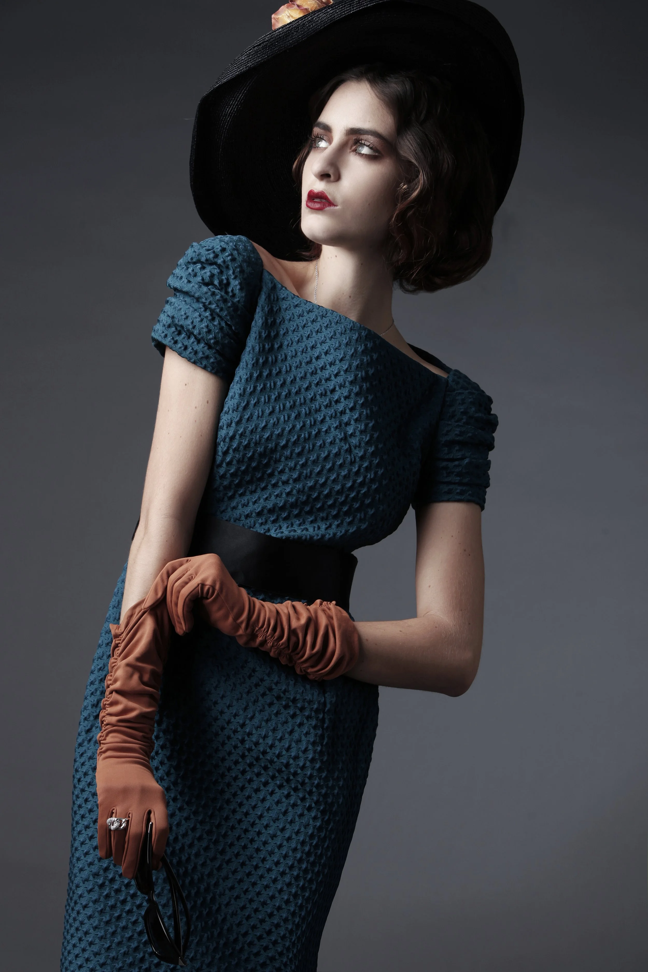 A woman with dark, curly hair wearing a large black hat, a blue textured dress, holding brown gloves, and posing against a plain dark background.