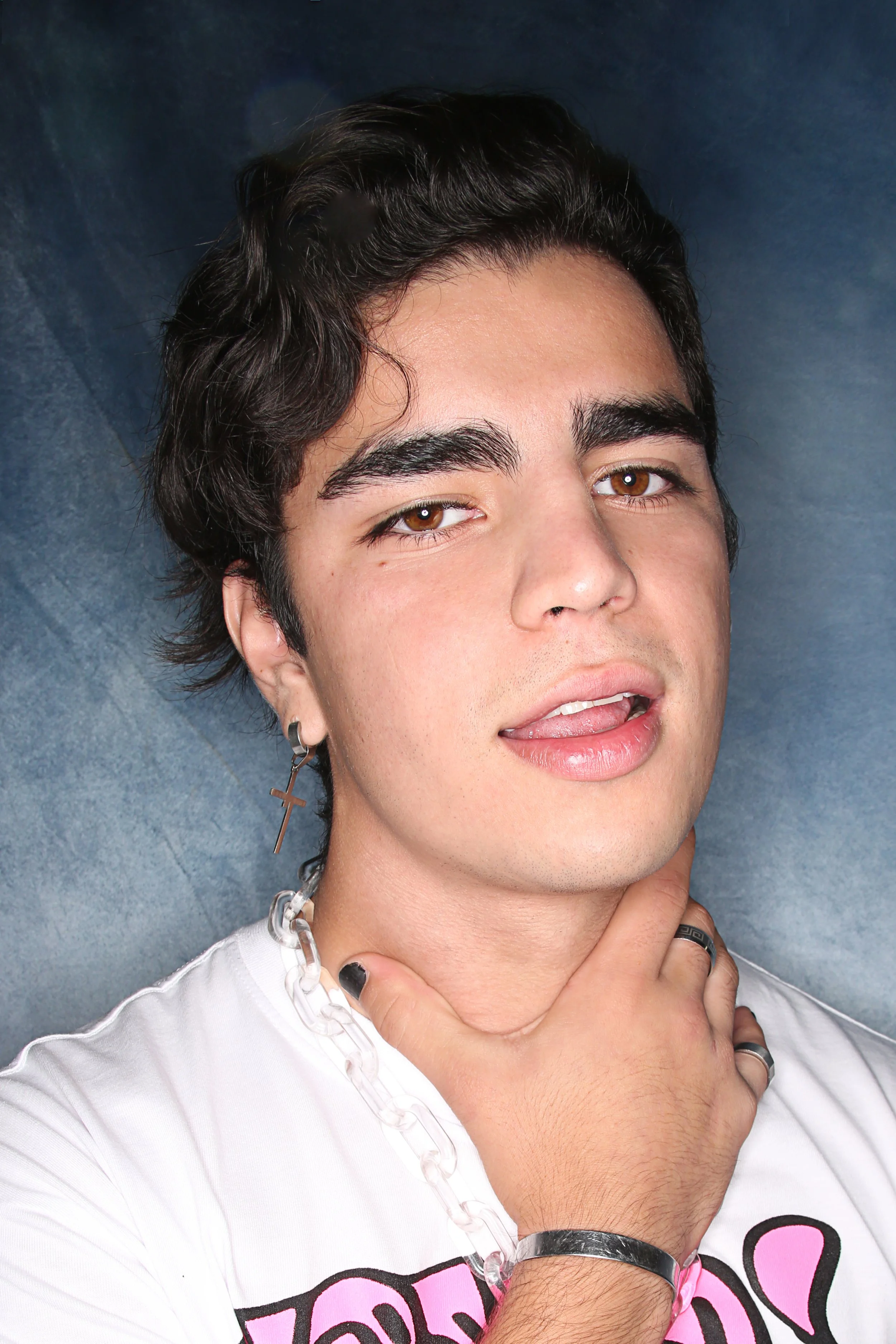 Close-up of a young man with dark hair and brown eyes, wearing earrings and accessories, touching his neck with slightly parted lips, against a dark blue background.