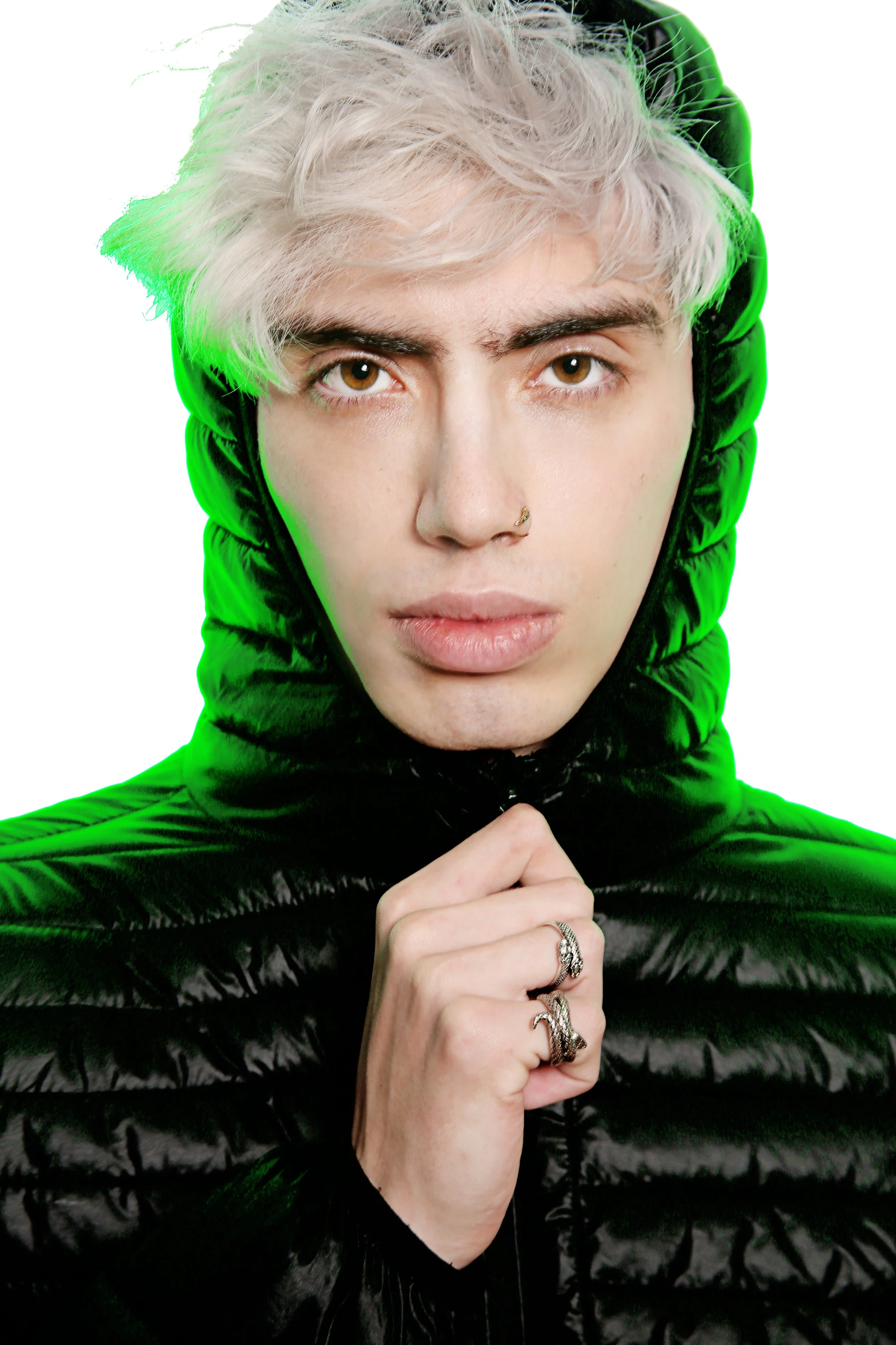 A person with light blonde, curly hair wearing a shiny black hooded jacket with green accents, holding up their hand with rings, against a white background.
