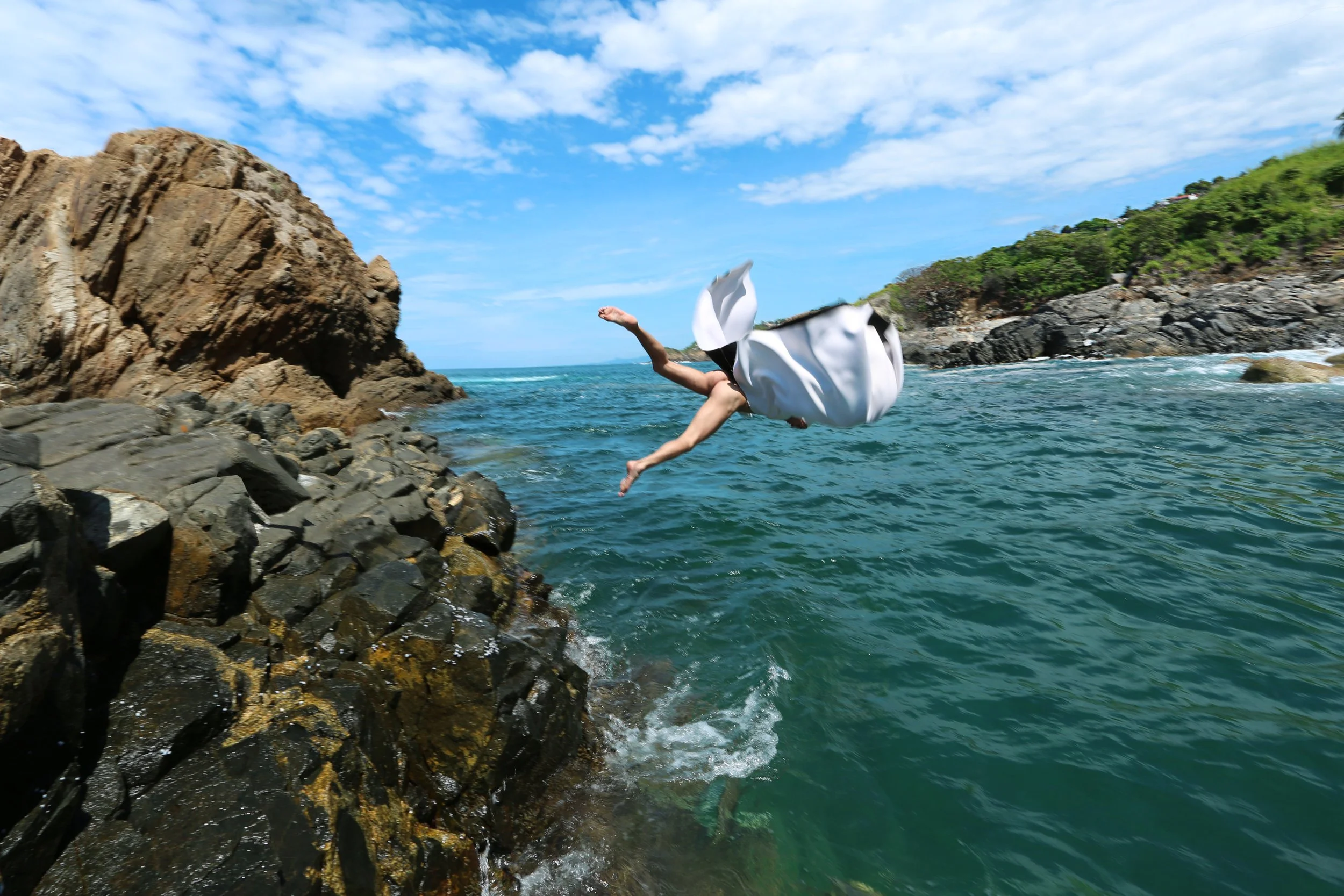 Person falling into the water from rocky shoreline with green hillside and blue sky in background.