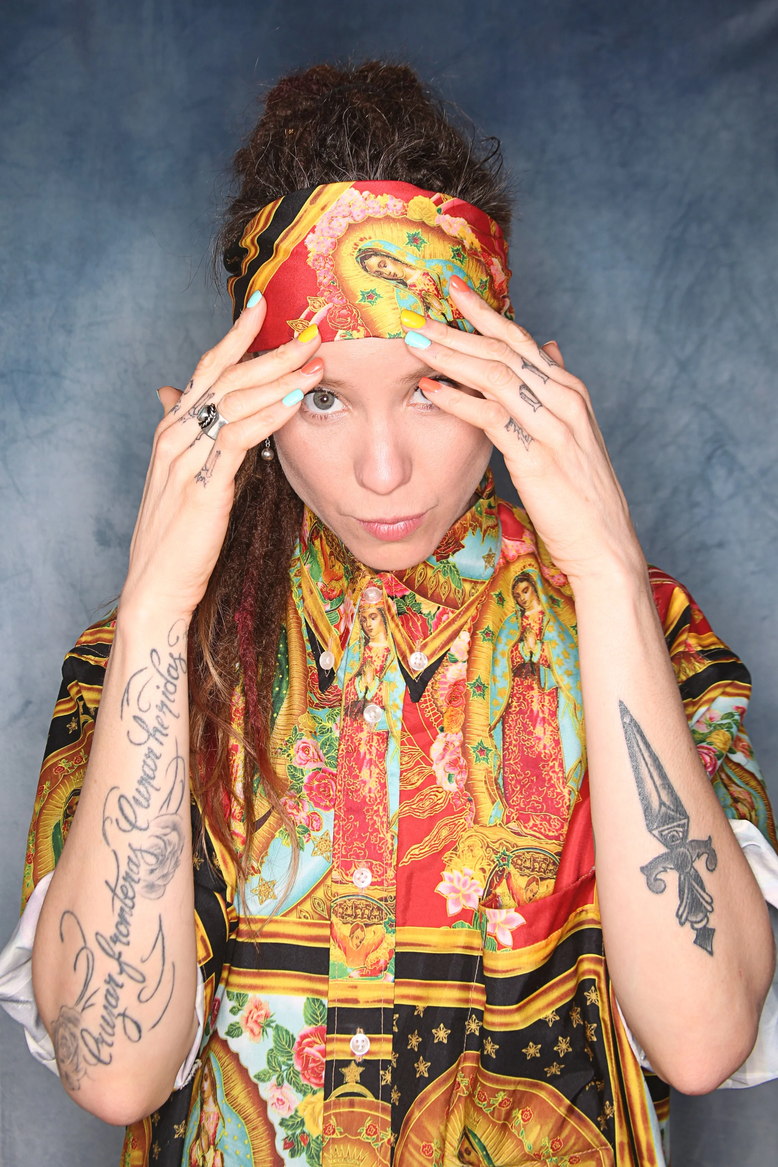 A woman with dark hair in dreadlocks wearing a colorful, patterned shirt and a red headscarf with religious images, holding her hands to her face.