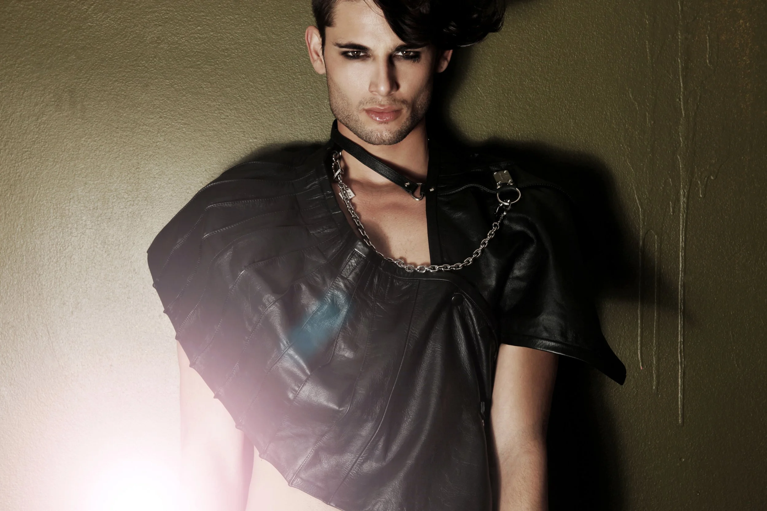 A young man with dark hair, wearing makeup, a black leather jacket, a choker, and a chain necklace, leaning against a green wall.