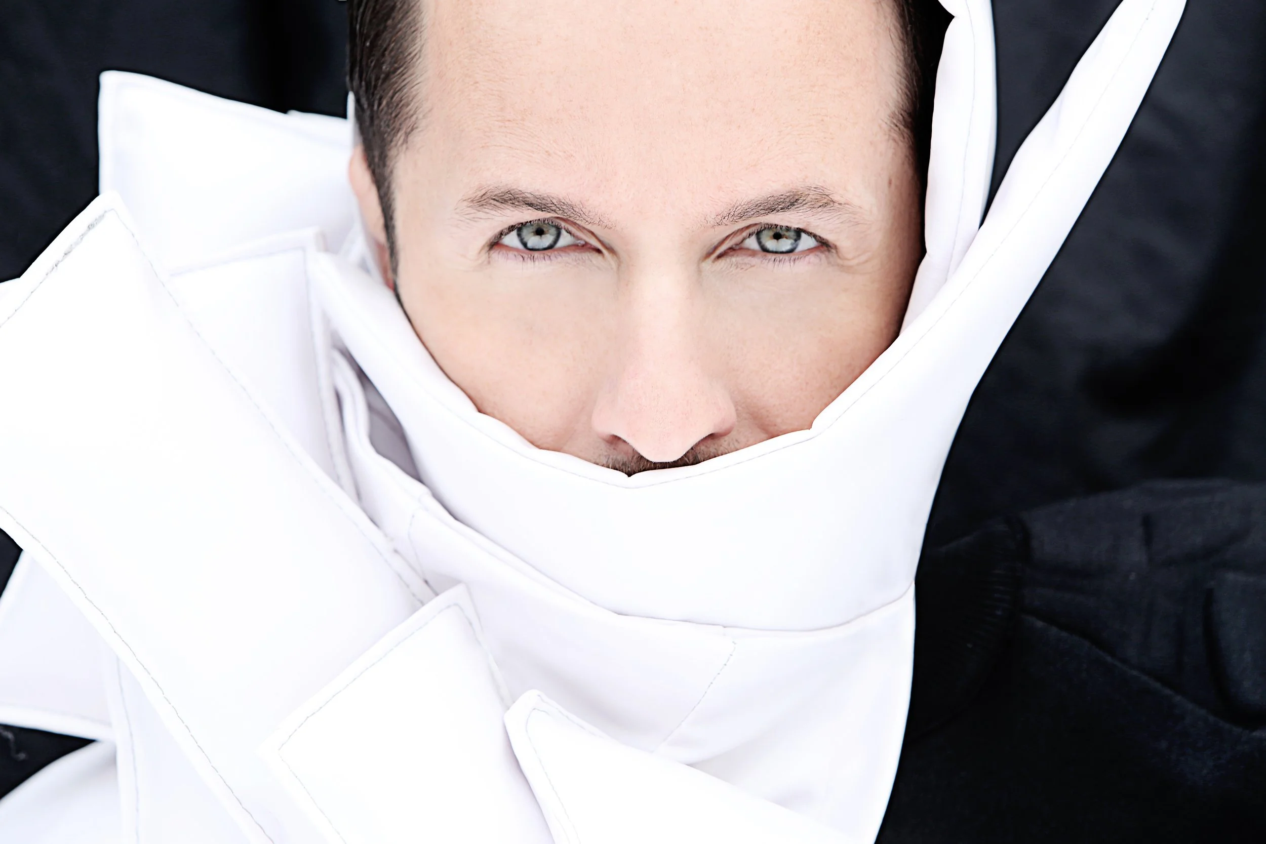 Close-up of a person's face with striking blue eyes, surrounded by white and black fabric, possibly a formal outfit.