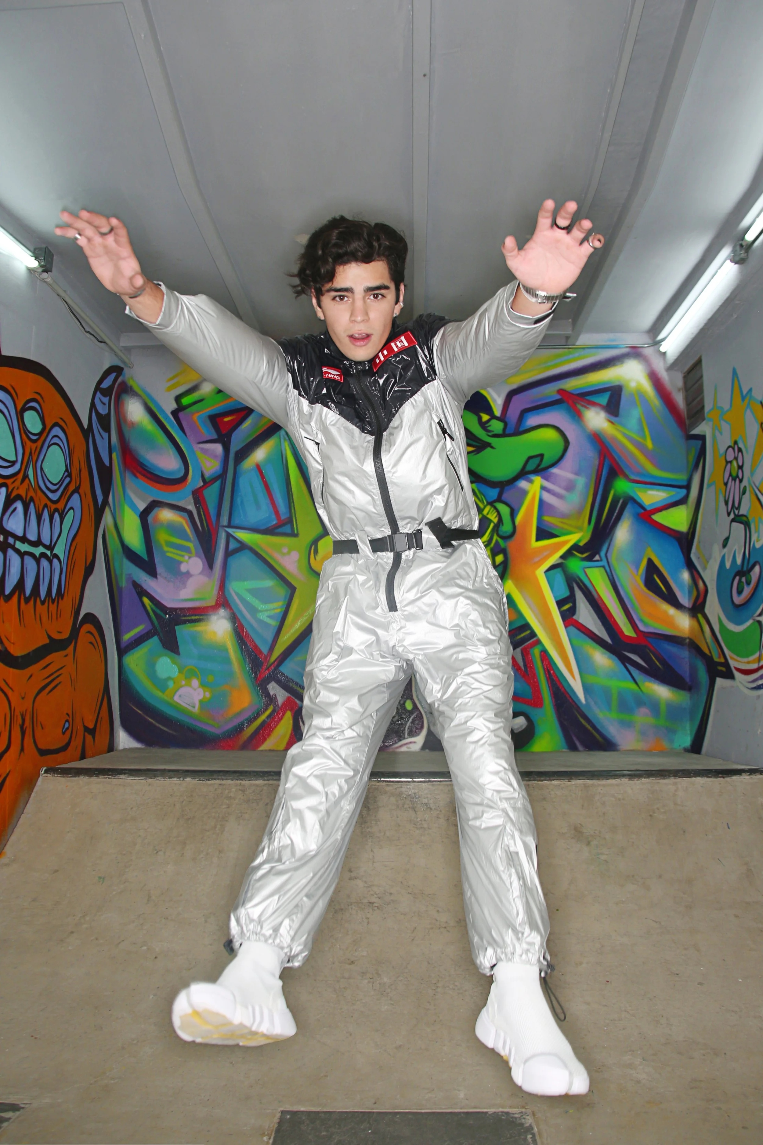 Young man in a metallic silver jacket and pants stands with arms raised in front of colorful graffiti art on the walls of an indoor skate park.