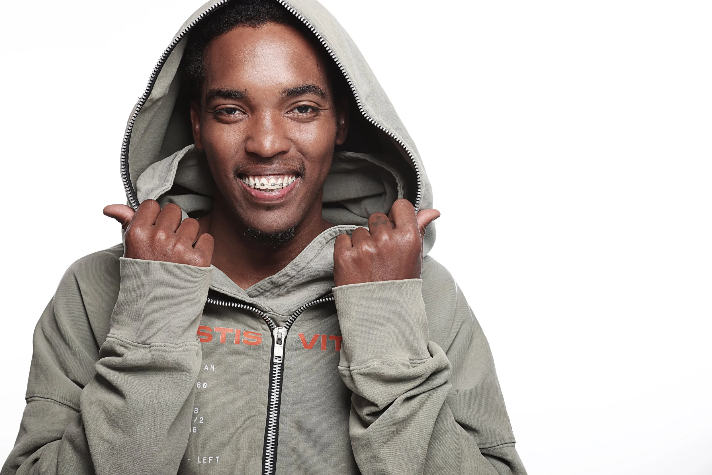A smiling person with dark skin wearing a gray hoodie and braces, holding the hoodie with both hands, against a white background.