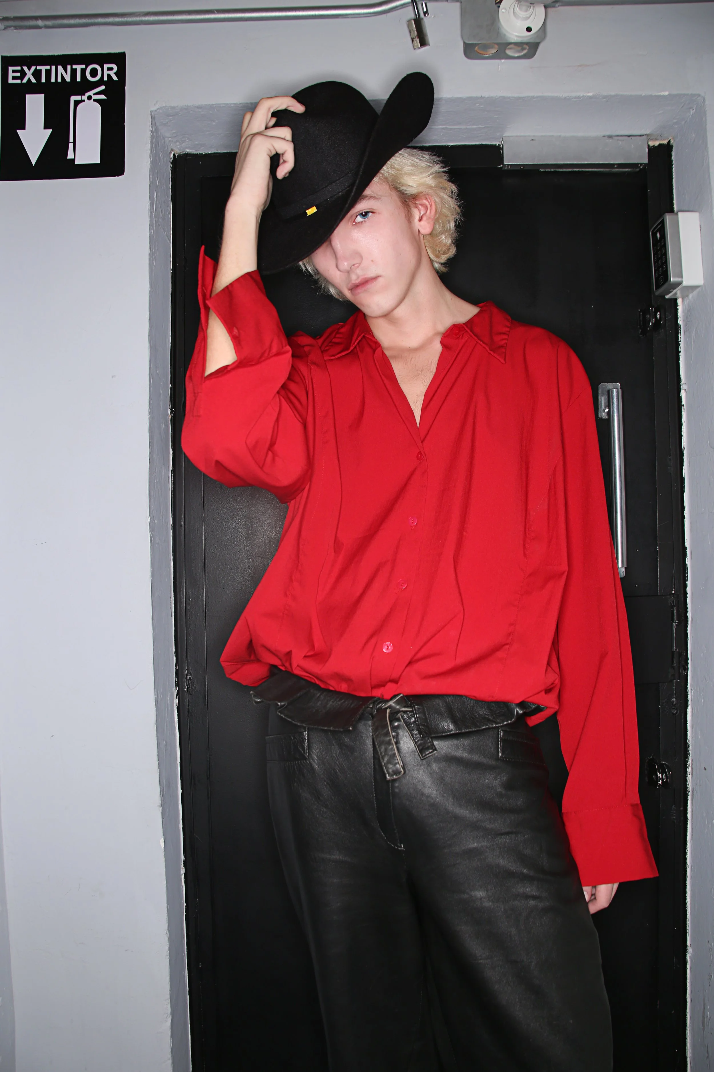 A person with blonde hair wearing a red shirt, black pants, and a black hat, leaning against a door frame in an indoor setting.