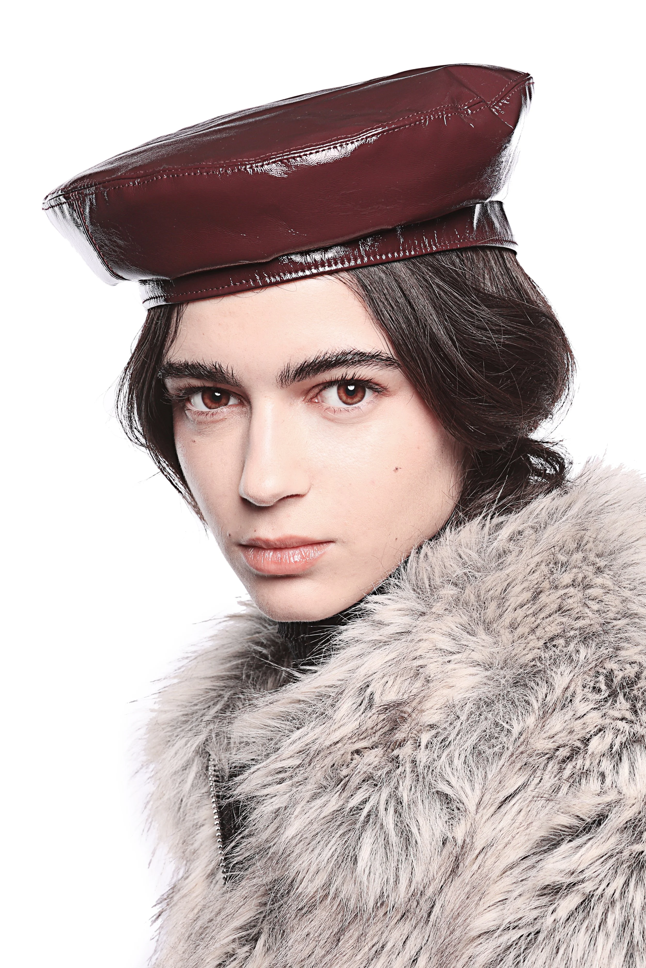 A young woman with dark hair and brown eyes wearing a dark red leather beret and a faux fur coat with gray tones, against a white background.