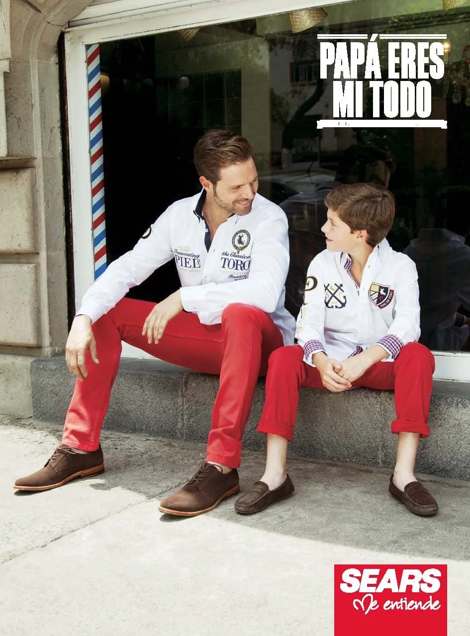 A father and son sitting on a sidewalk outside a shop window, smiling and looking at each other. Both are wearing white shirts with logos and red pants, with the father’s shoes being brown and the son's shoes being dark brown loafers. The window feat