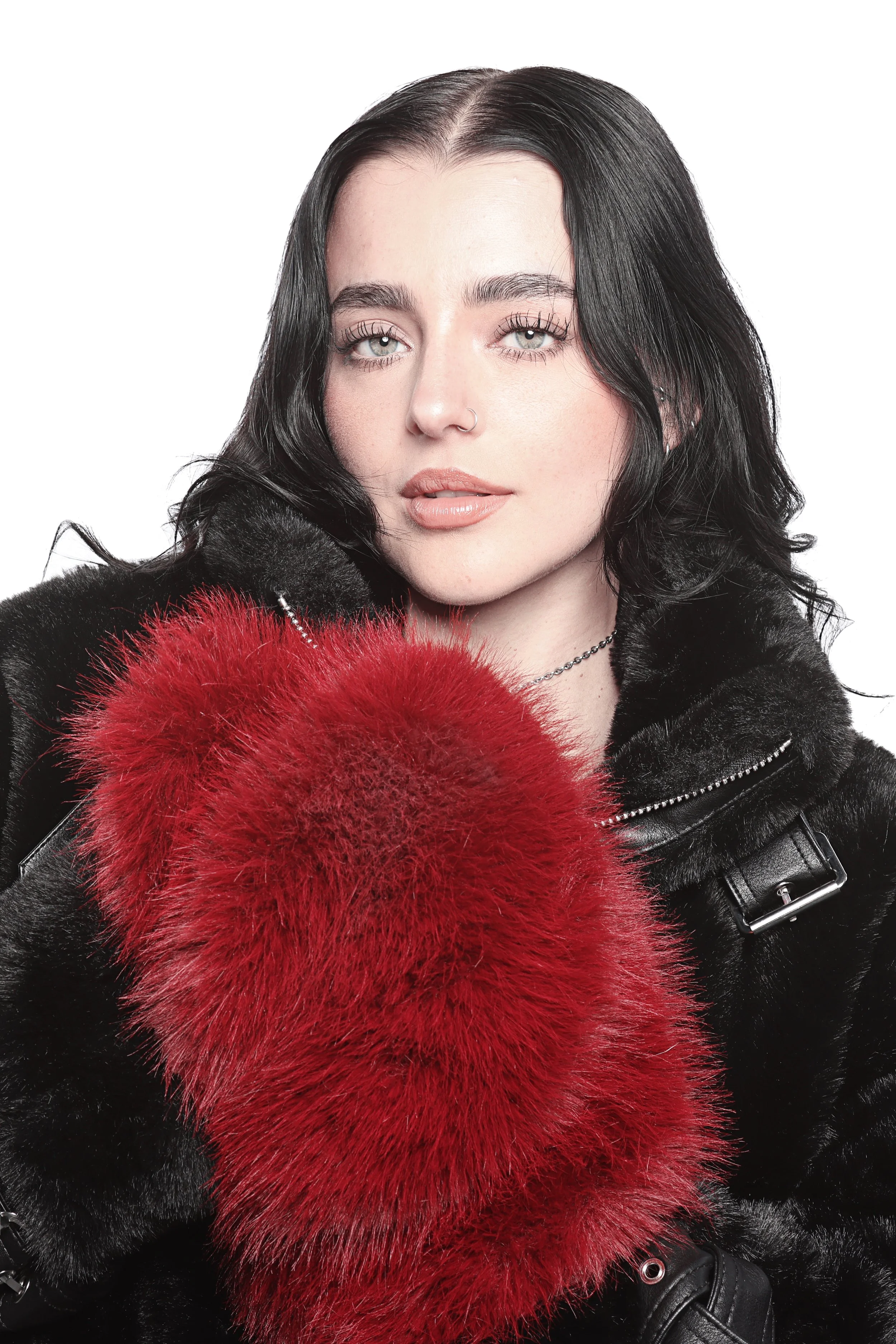 A woman with long black hair, light blue eyes, and a nose ring, wearing a black leather jacket with buckles, holding a red and black faux fur stole against a white background.