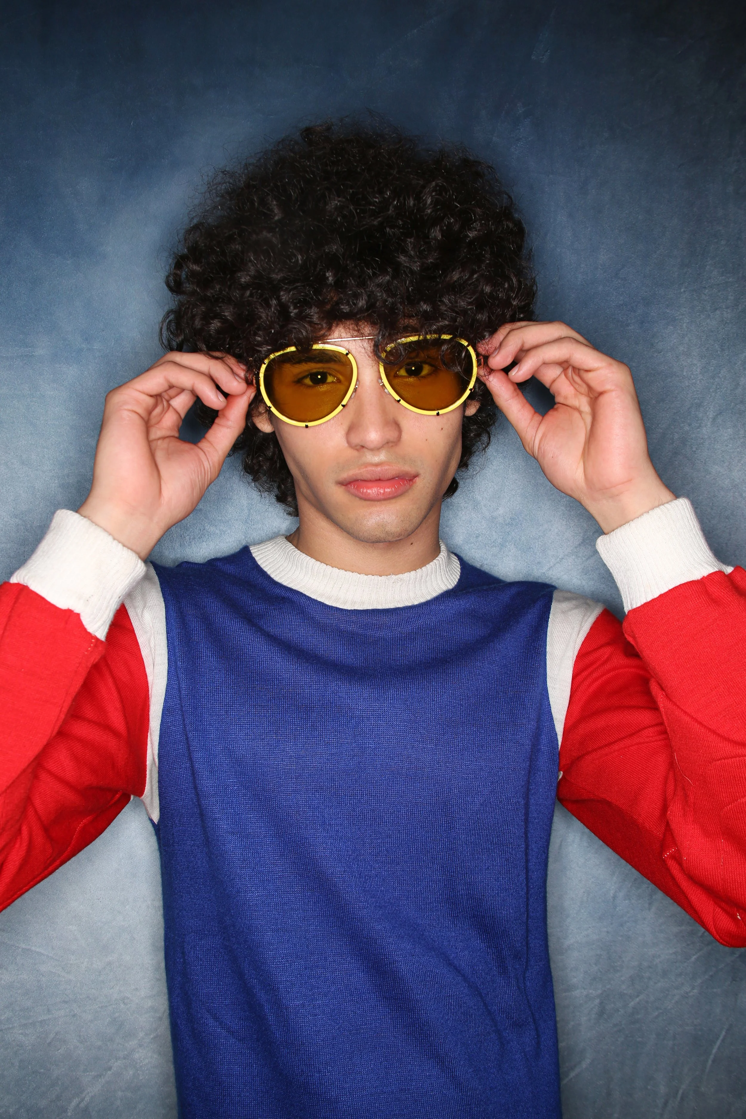 A person with curly dark hair wearing yellow-tinted sunglasses, a multicolored long-sleeve shirt with red, white, and blue sections, and lying on a gray fabric surface.