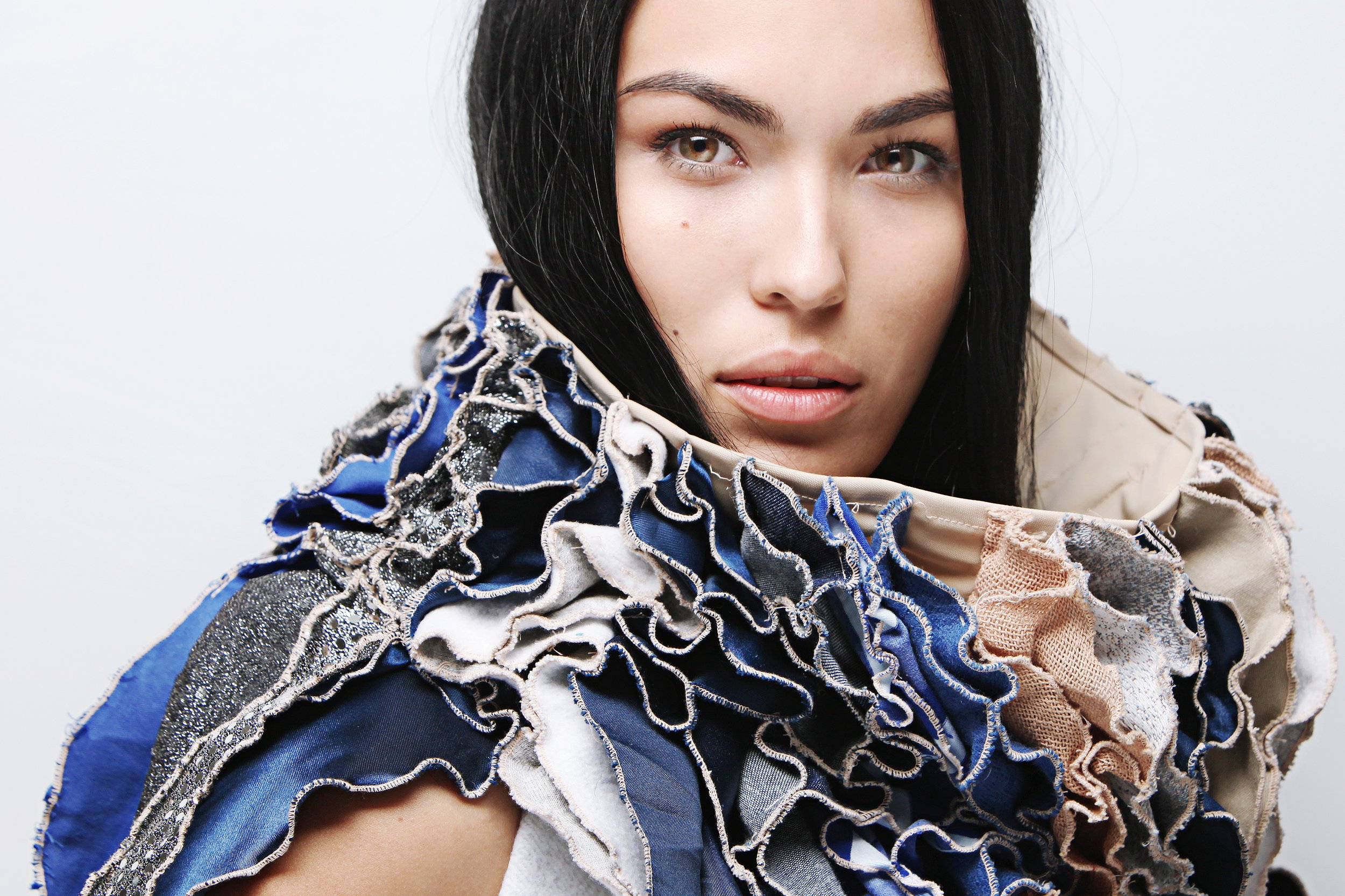 Close-up portrait of a woman with dark hair and brown eyes, wearing a multicolored, textured fabric jacket with ruffled details.