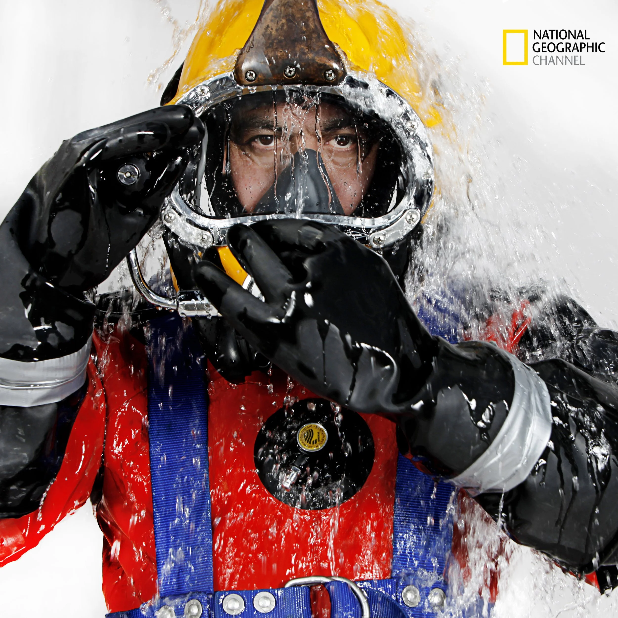 A person wearing a yellow helmet, black gloves, and a red jacket, looking directly at the camera with water running down their face, holding the visor of the helmet. The background is white. The National Geographic Channel logo is in the top right co