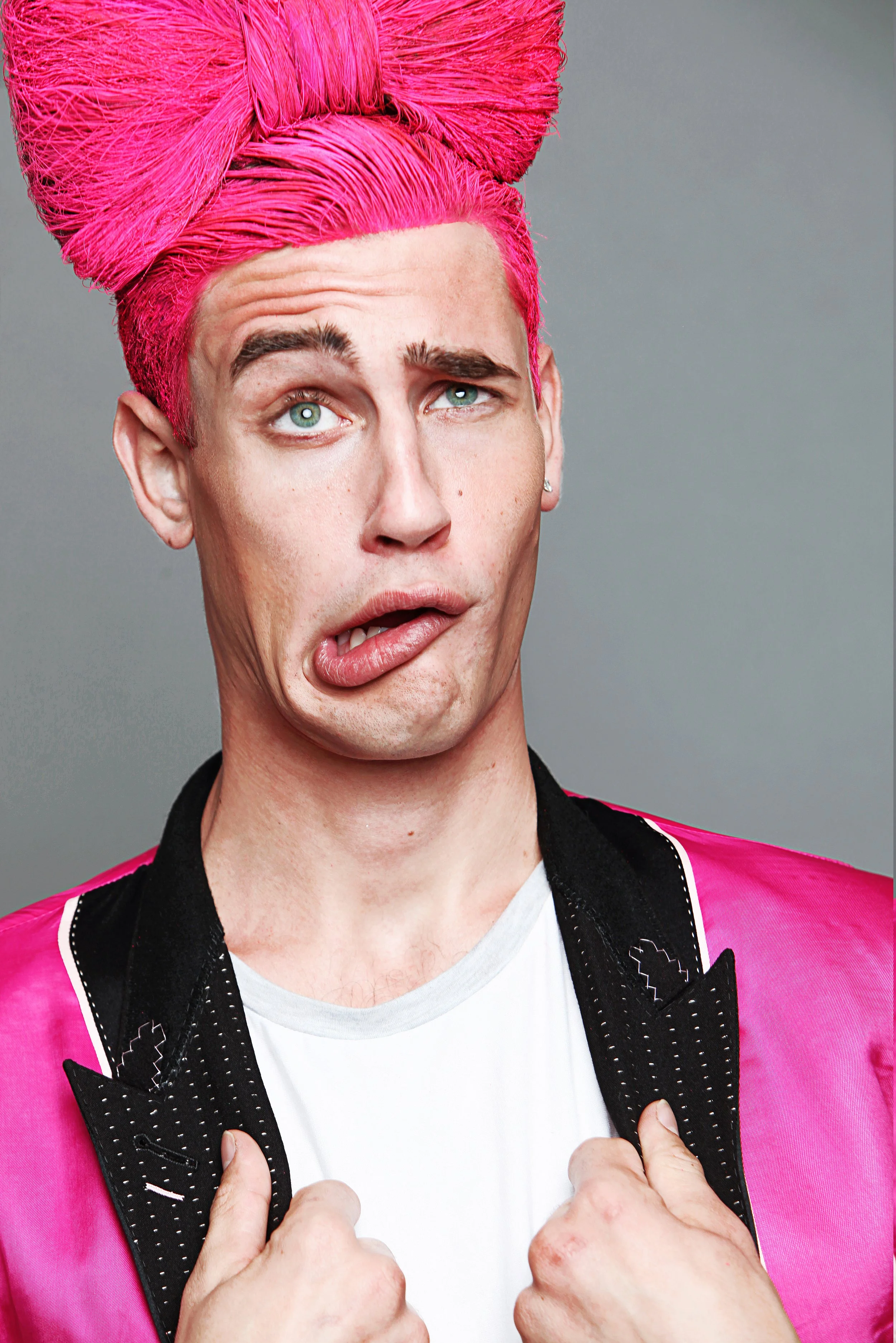 Person with bright pink hair styled in an updo, making a humorous facial expression with tongue slightly out, holding the lapel of a pink jacket with black detailing, wearing a white shirt underneath, in front of a plain gray background.